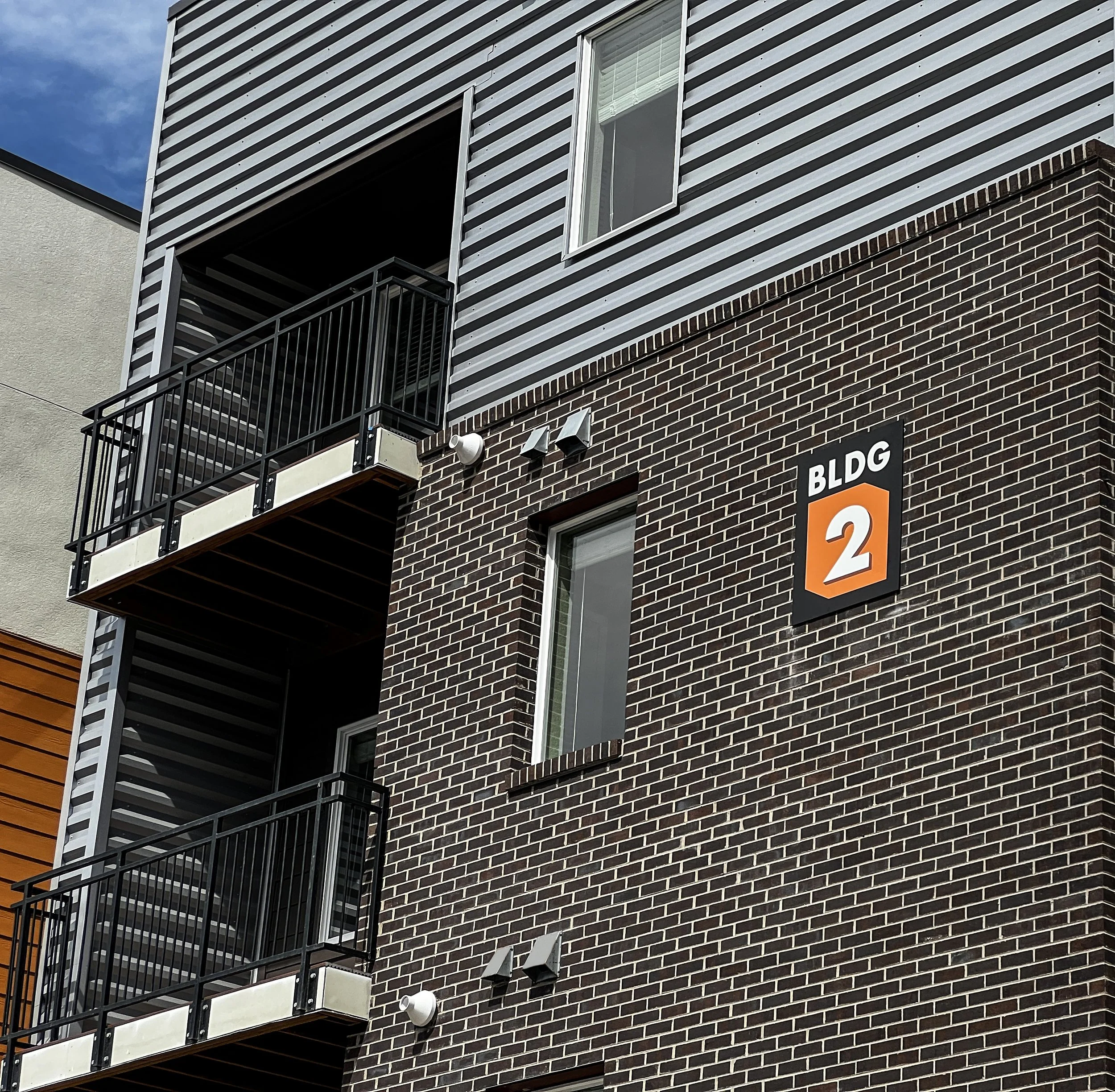 Close-up of a modern apartment building exterior with dark brick wall, two balconies with black metal railings, a white framed window, and an orange and black building sign labeled 'BLDG 2'.