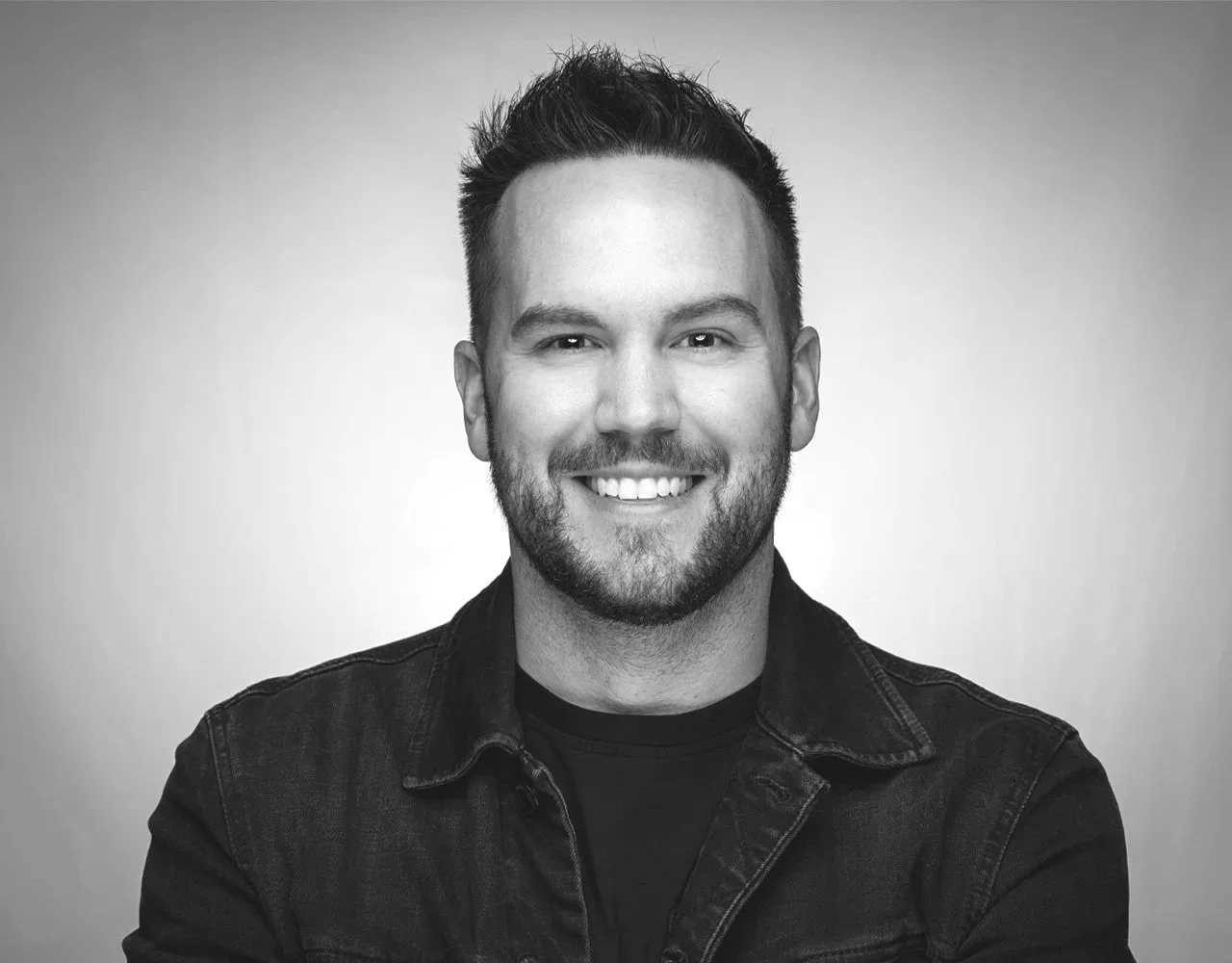 Black and white portrait of a young man with short hair, a beard, smiling, wearing a dark denim jacket, against a plain background.
