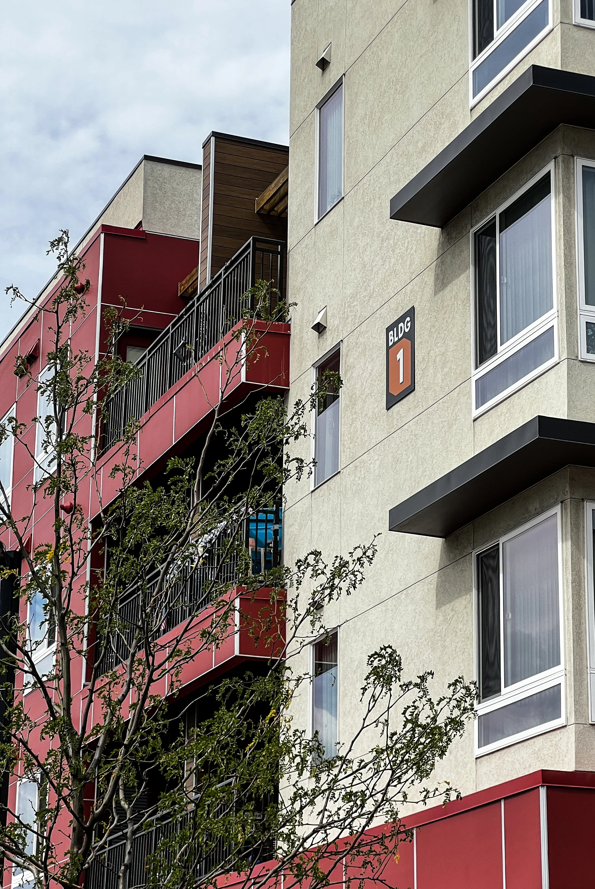 Exterior view of a multi-story residential building with red balconies and a yellow facade, with tree branches in the foreground and a sign that reads 'BLDG 1' on the building.