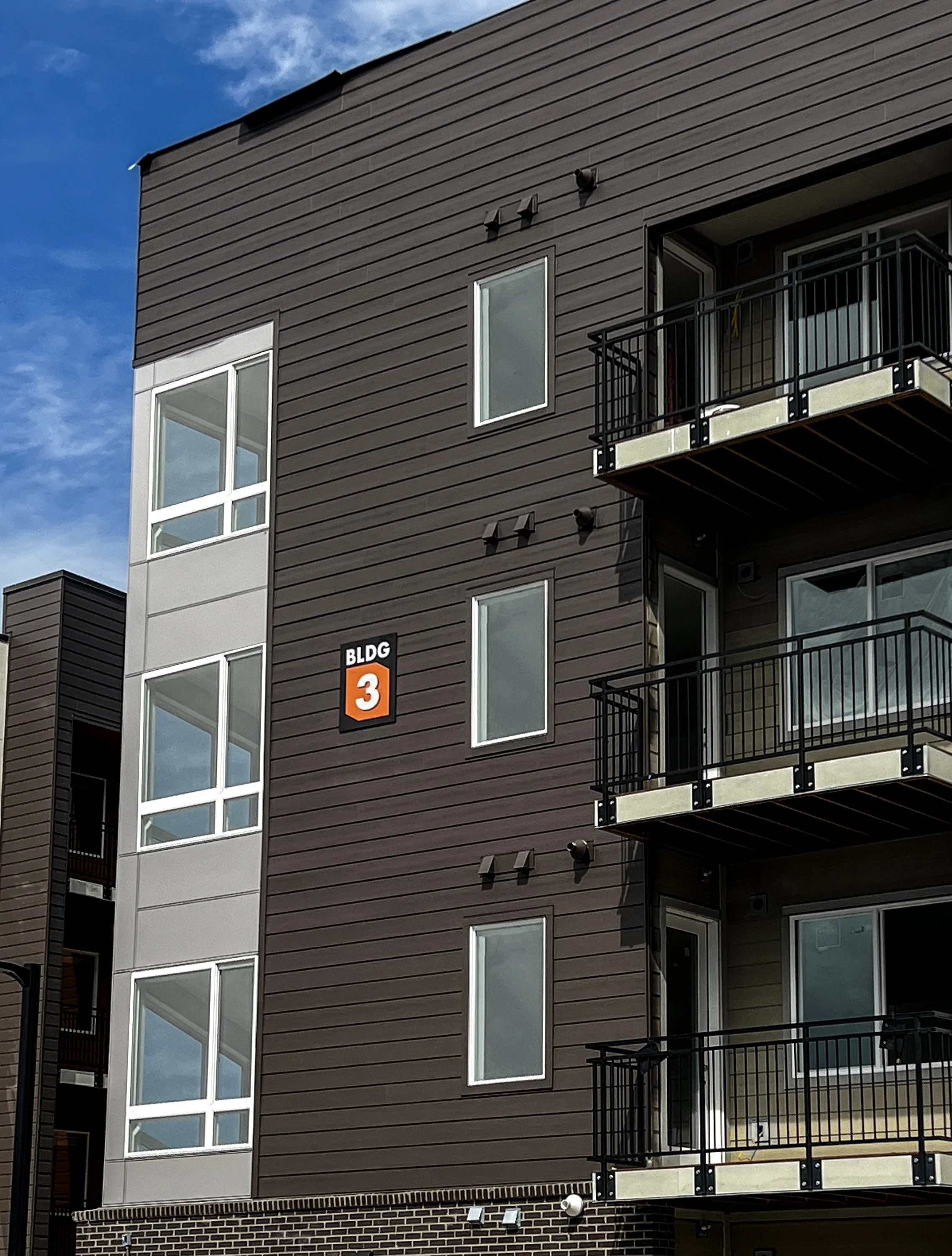 Building ID sign modern multi-story apartment building with brown and gray exterior siding, balconies with black railings, and a sign reading 'BLDG 3' on the wall.