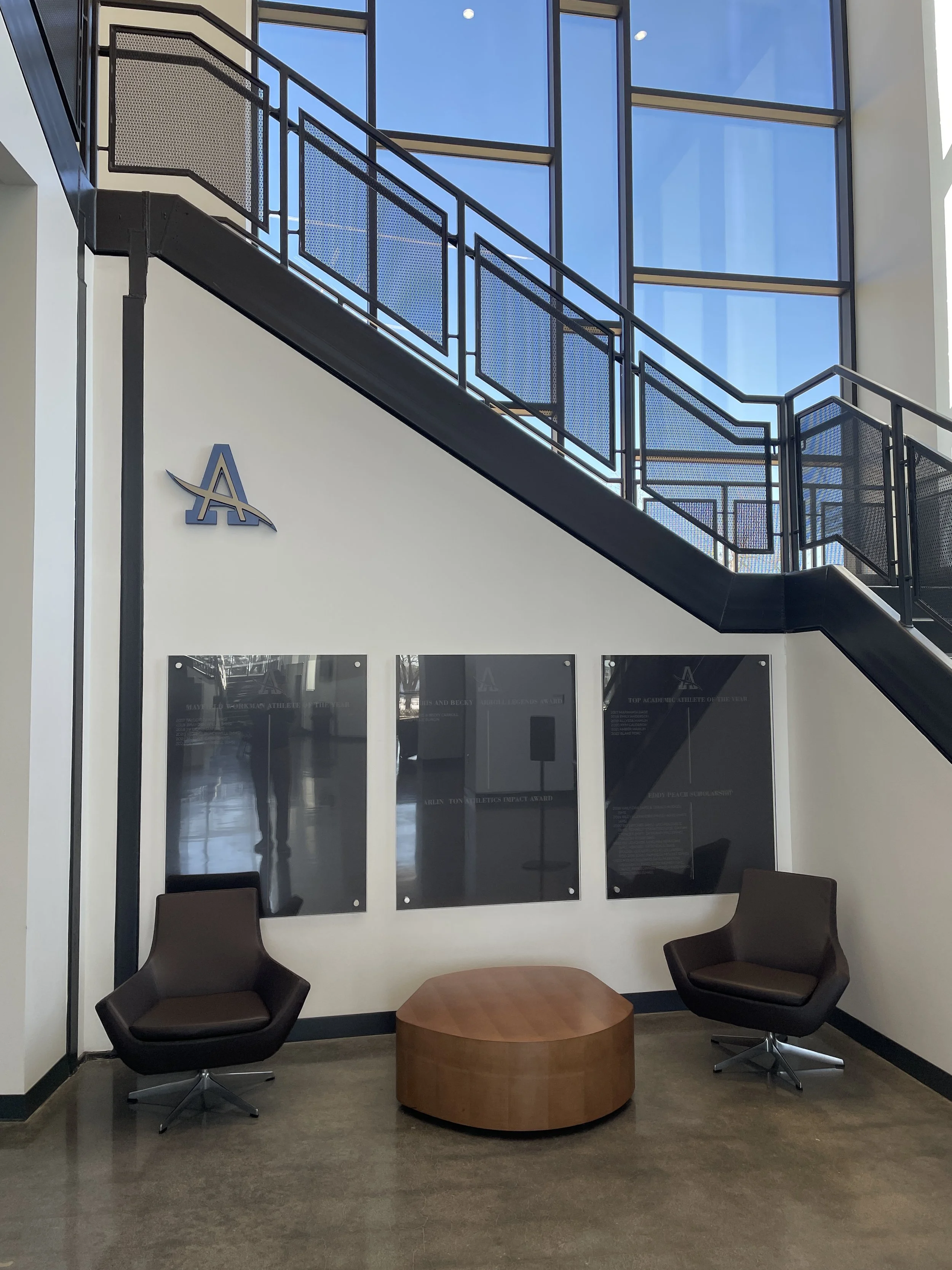 Interior of a modern building lobby with two brown chairs, a wooden coffee table, black plaques on the wall, and a black metal staircase with large windows in the background.