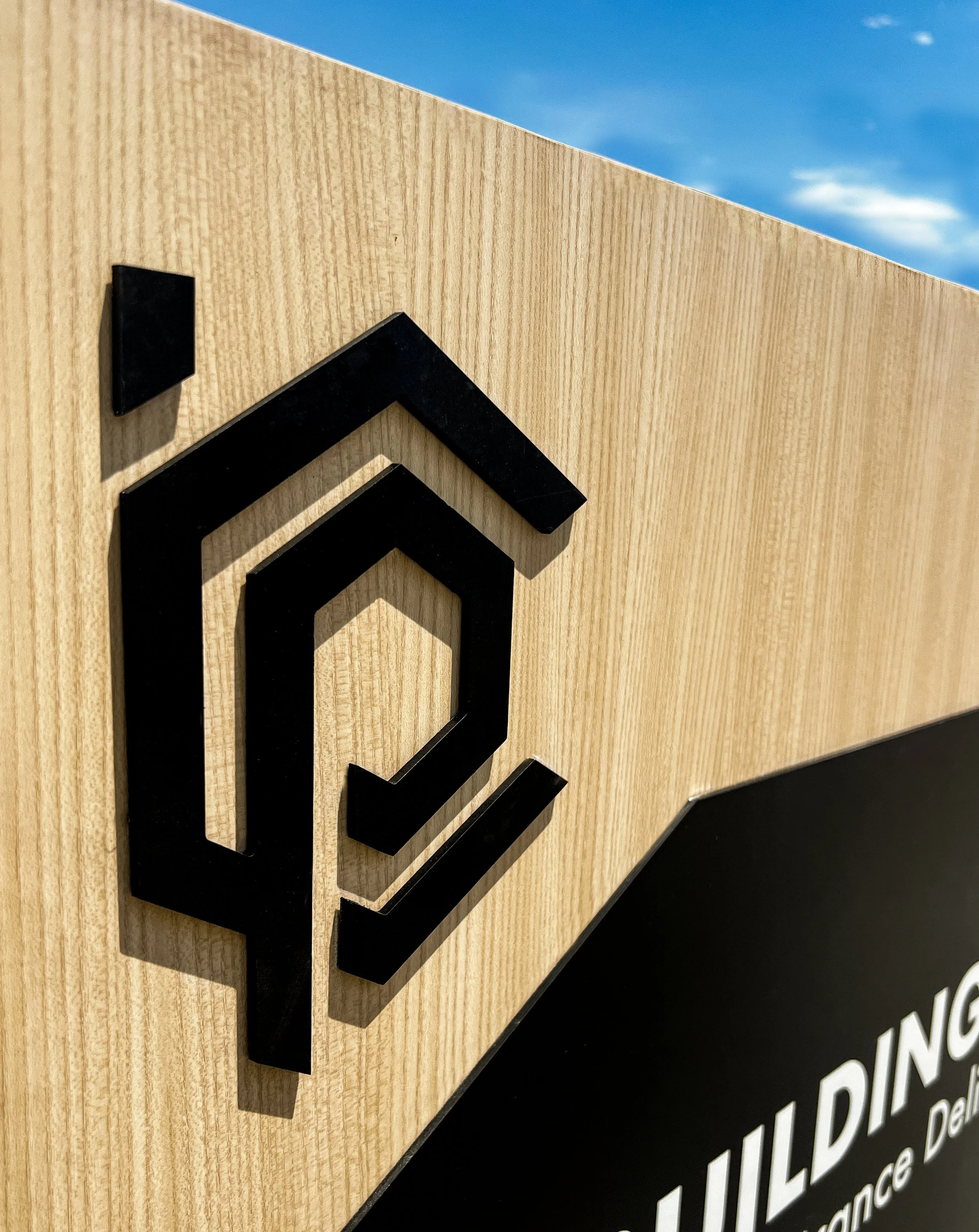 Close-up of a wooden sign with a three-dimensional black logo and partial text, against a blue sky background.