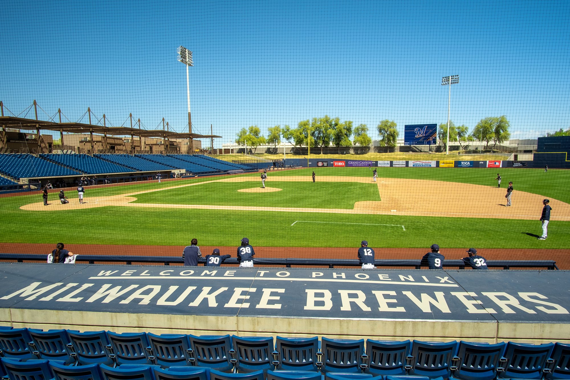 View of a baseball stadium with players on the field, synthetic netting in the foreground, and an empty seating area with blue seats. The field has players warming up and a sign that reads 'Welcome to Phoenix Milwaukee Brewers.'