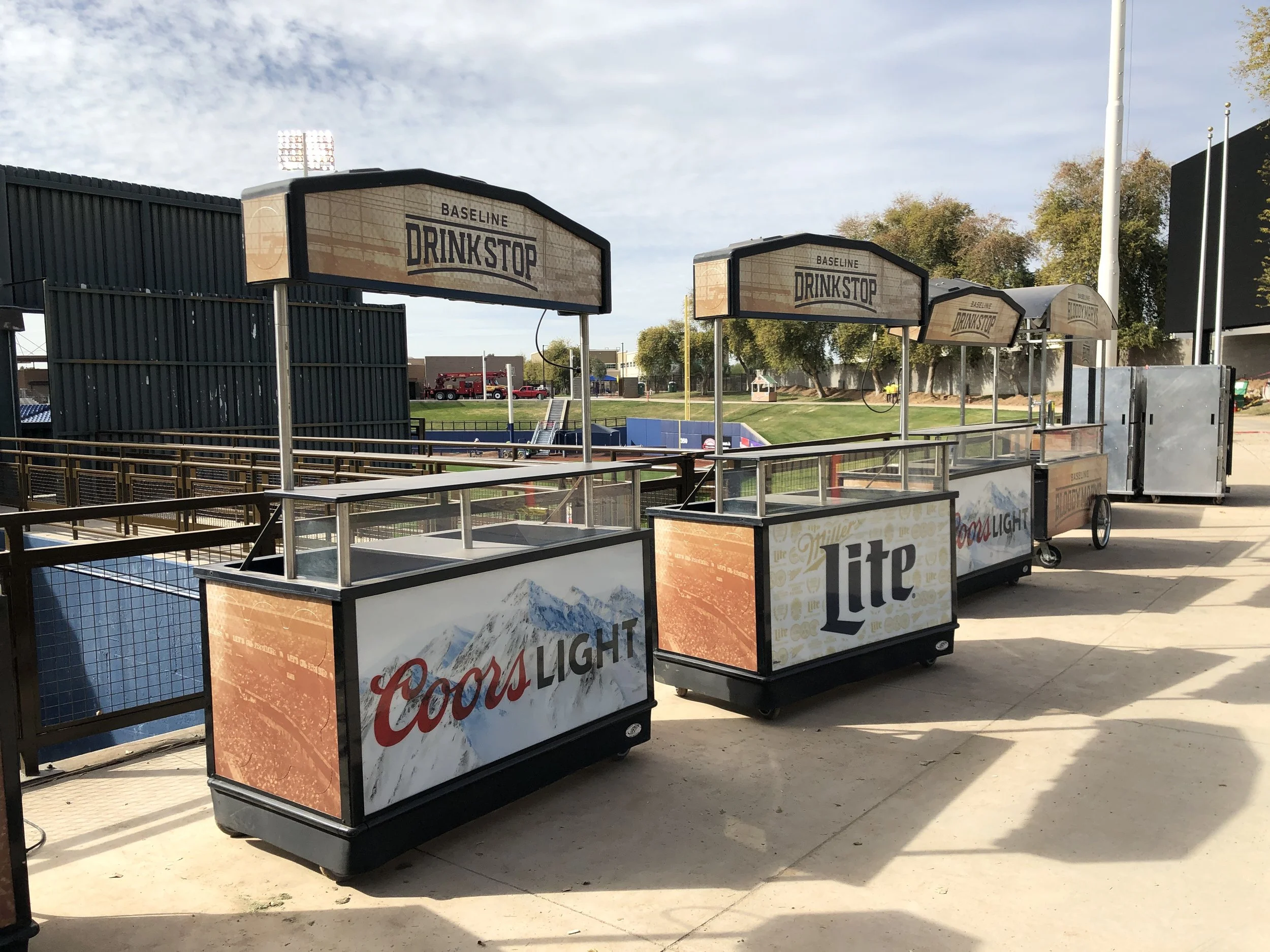 Set up for outdoor beer or food stand with Drink Stop and Coors Light branding, several service counters, and a grassy park area in the background for the MLB Milwaukee Brewers.