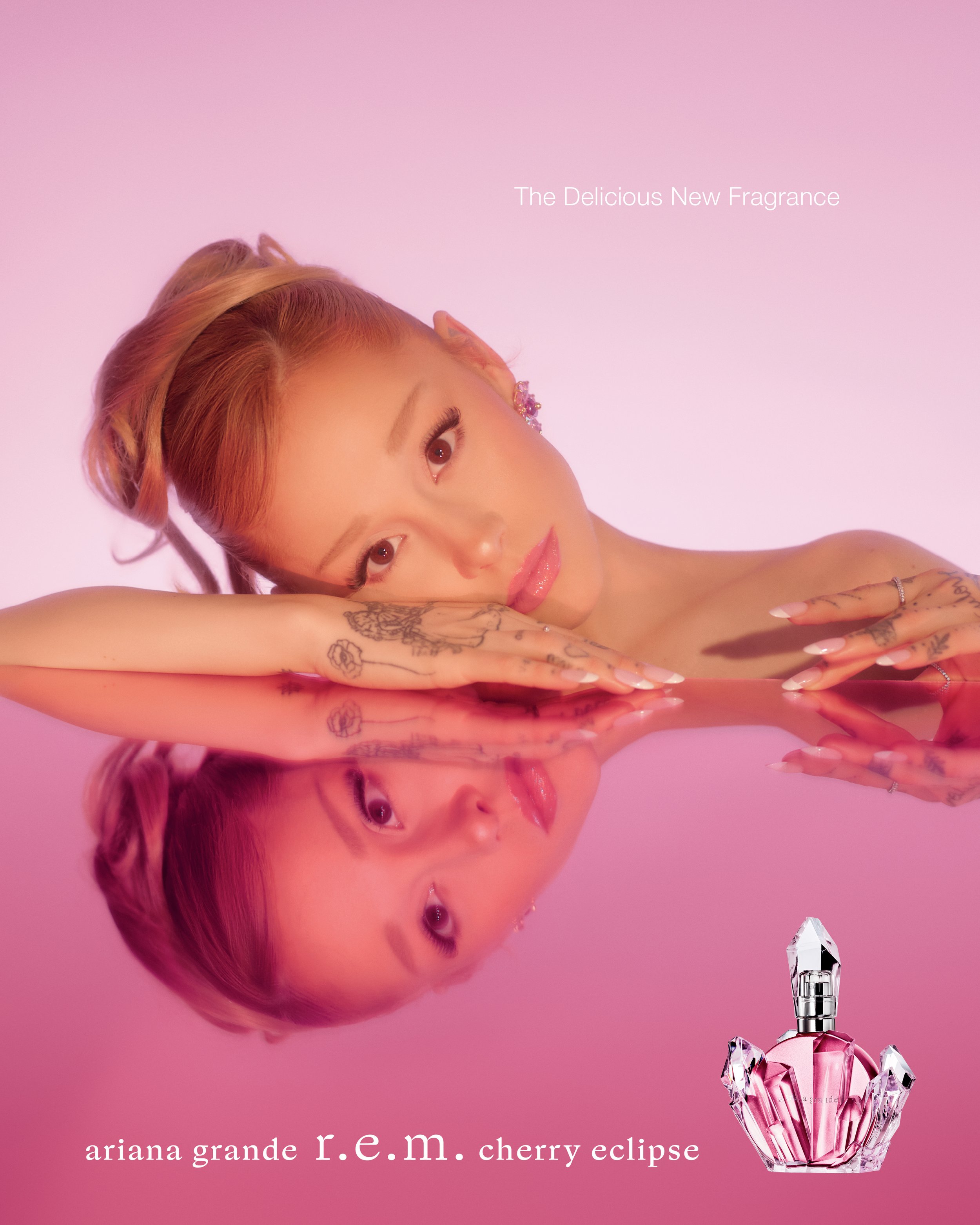 A woman with light skin, pink lipstick, and tattoos on her hands rests her head on her folded arms, reflected on a glossy surface with a pink background. Text on the image promotes Ariana Grande's fragrance 'R.E.M. Cherry Eclipse' and describes it as