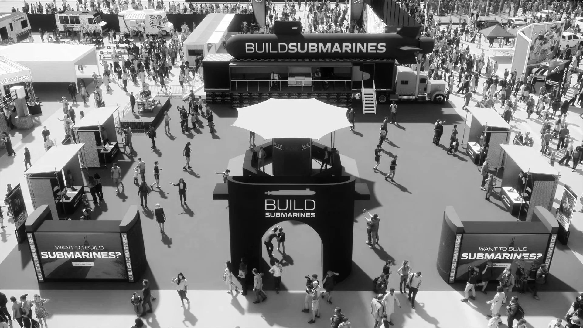A busy outdoor event with many people walking around, featuring a central booth with a canopy and promotional signage for building submarines, and a large truck behind it with a missile display on top that reads 'BUILD SUBMARINES.'