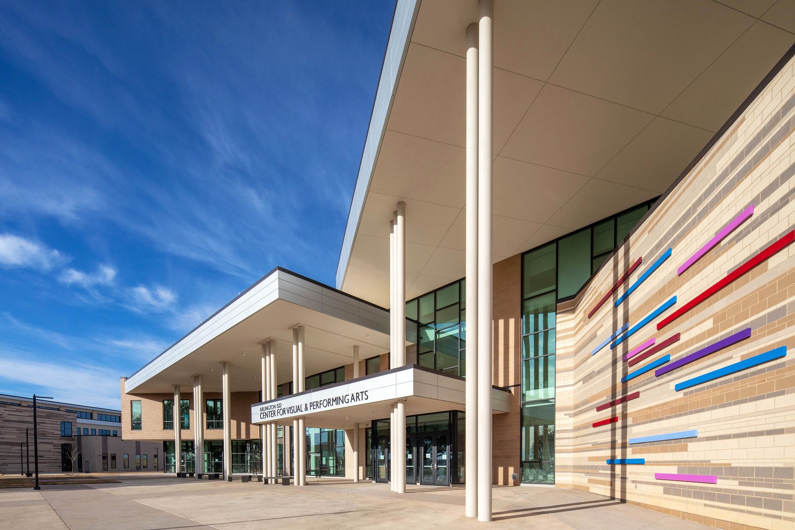 Modern building with large glass windows, white columns, and colorful metal art on the brick wall in front of a blue sky.