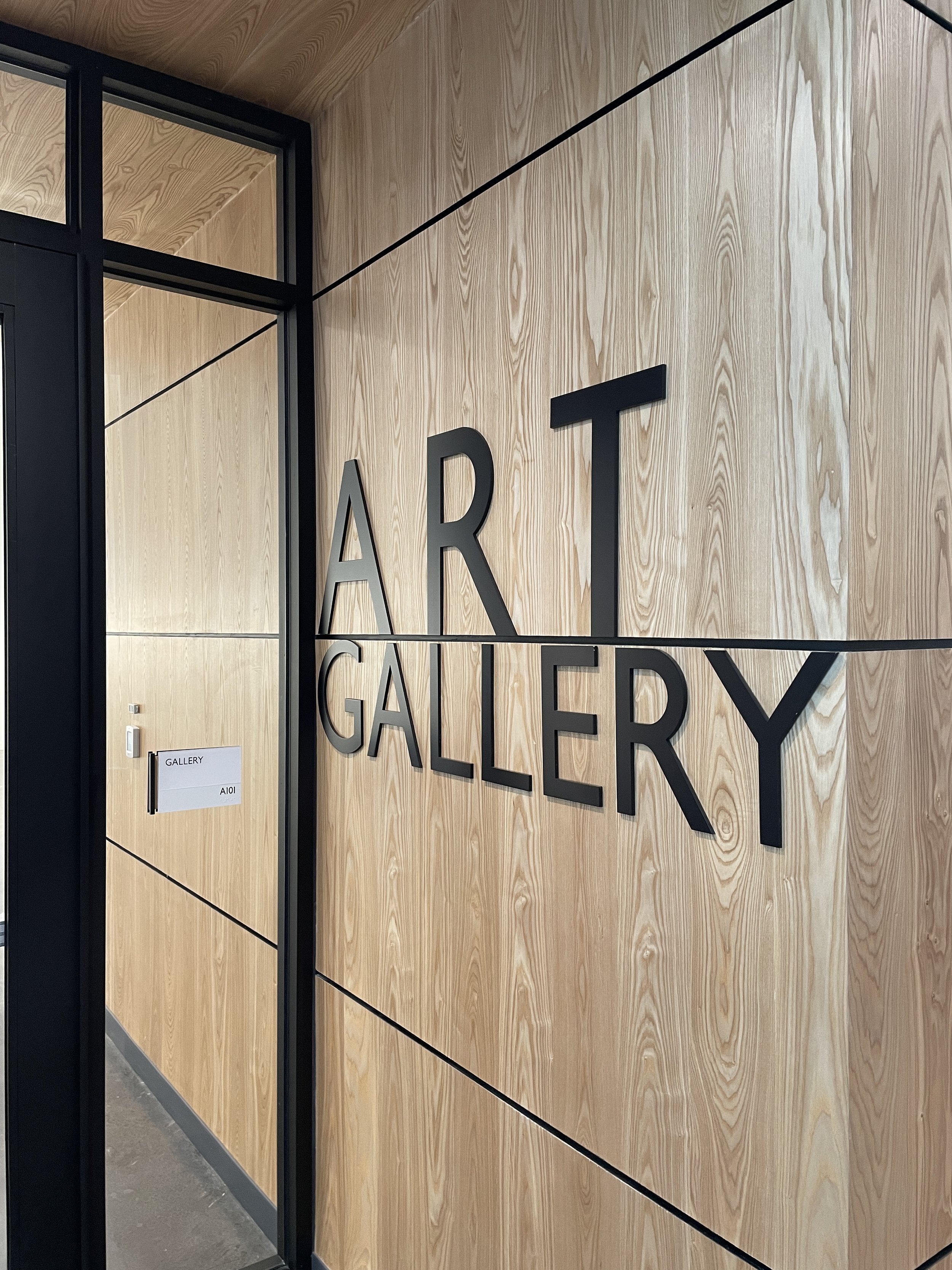 Interior of an art gallery with a wooden wall displaying black 3D letters spelling 'ART GALLERY' and a door with a sign reading 'GALLERY AI'.