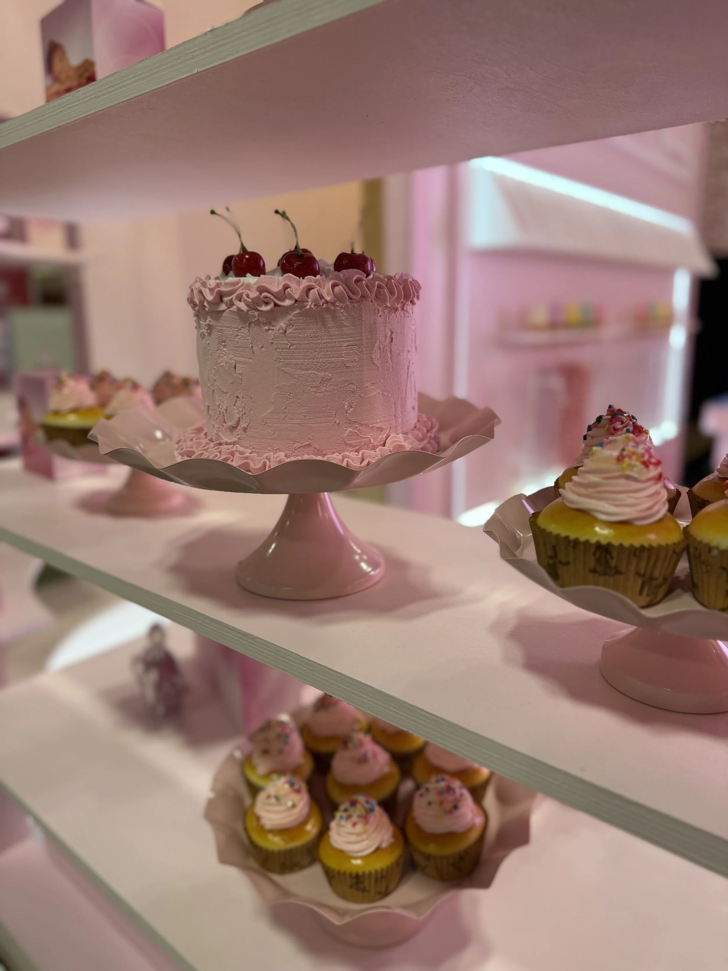 Pink frosted cake topped with three cherries on a pink cake stand, surrounded by pink frosted cupcakes with sprinkles, displayed on a white shelf with a pink background.