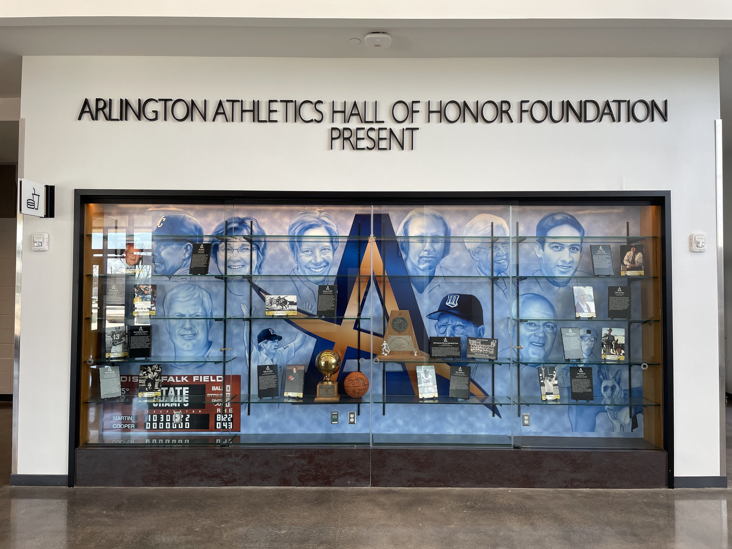 Display case featuring portraits of notable figures, sports memorabilia, and a large logo of the Dallas Mavericks basketball team, with a mural inside the Arlington Athletics Hall of Honor Foundation.