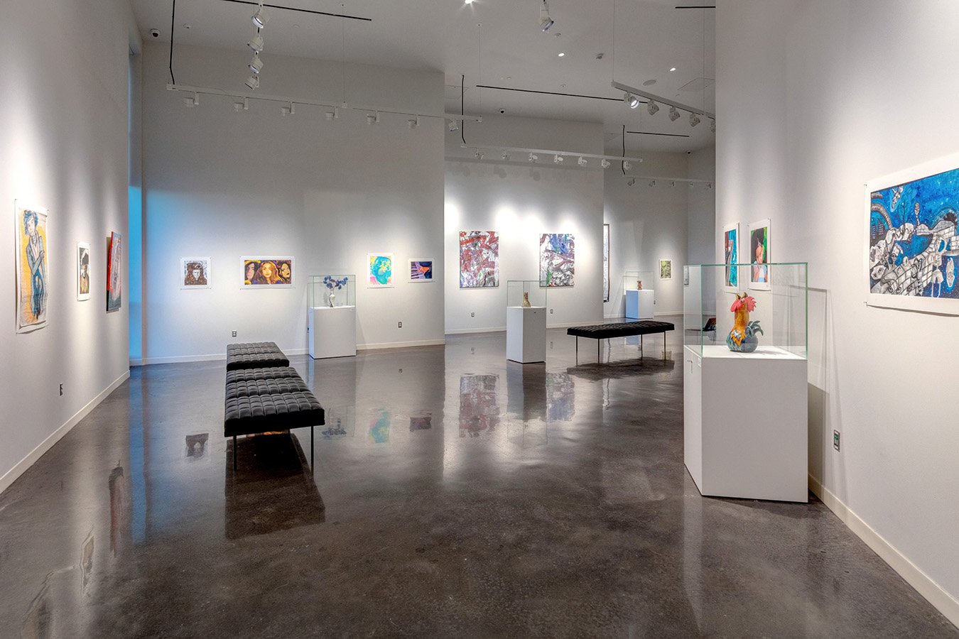 An art gallery with paintings and sculptures on display, white walls, polished concrete floor, black cushioned benches, and track lighting overhead.