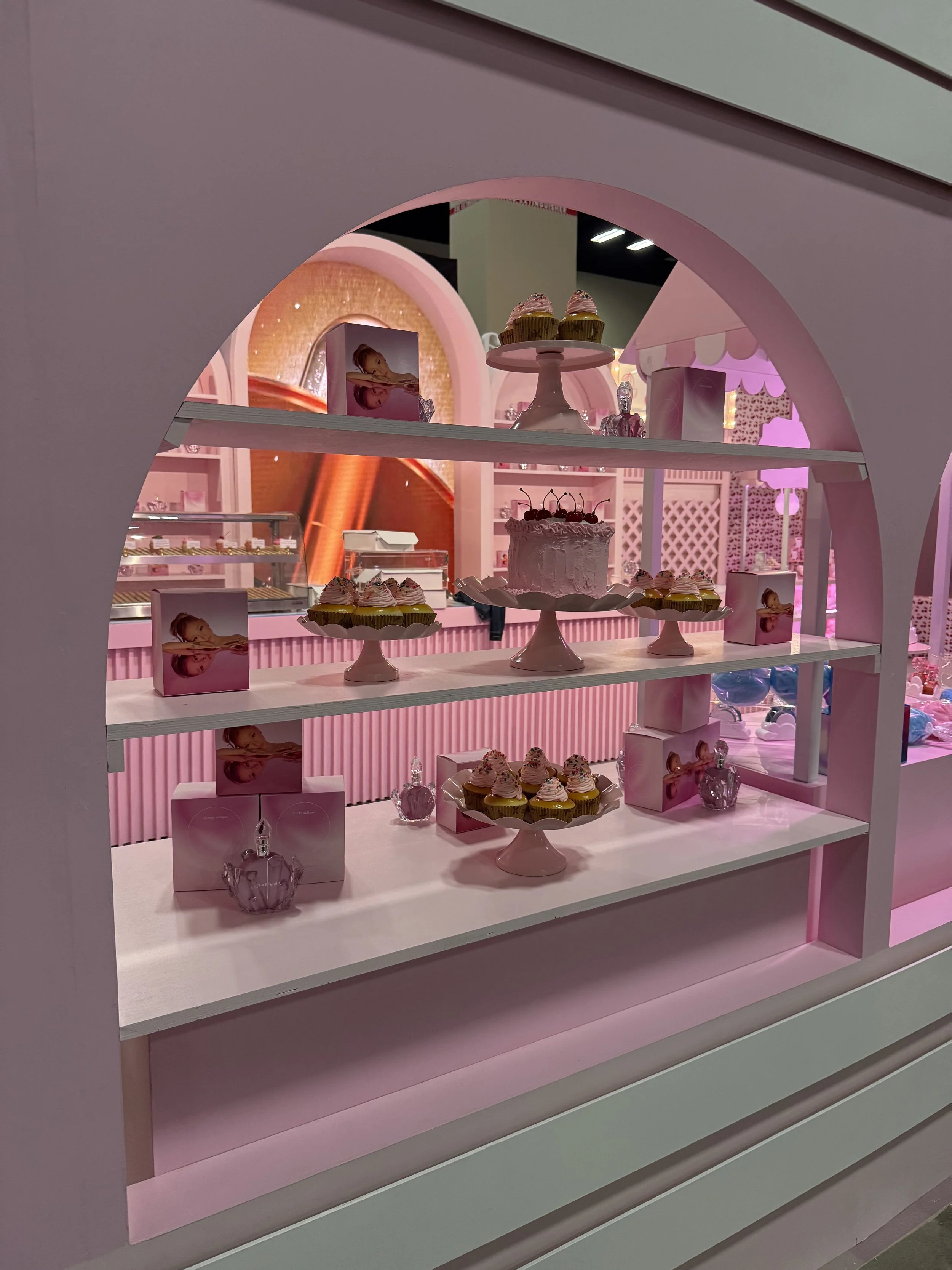 Display of cupcakes and decorative items in a pastel pink-themed bakery or dessert shop, viewed through a window with an arch shape.