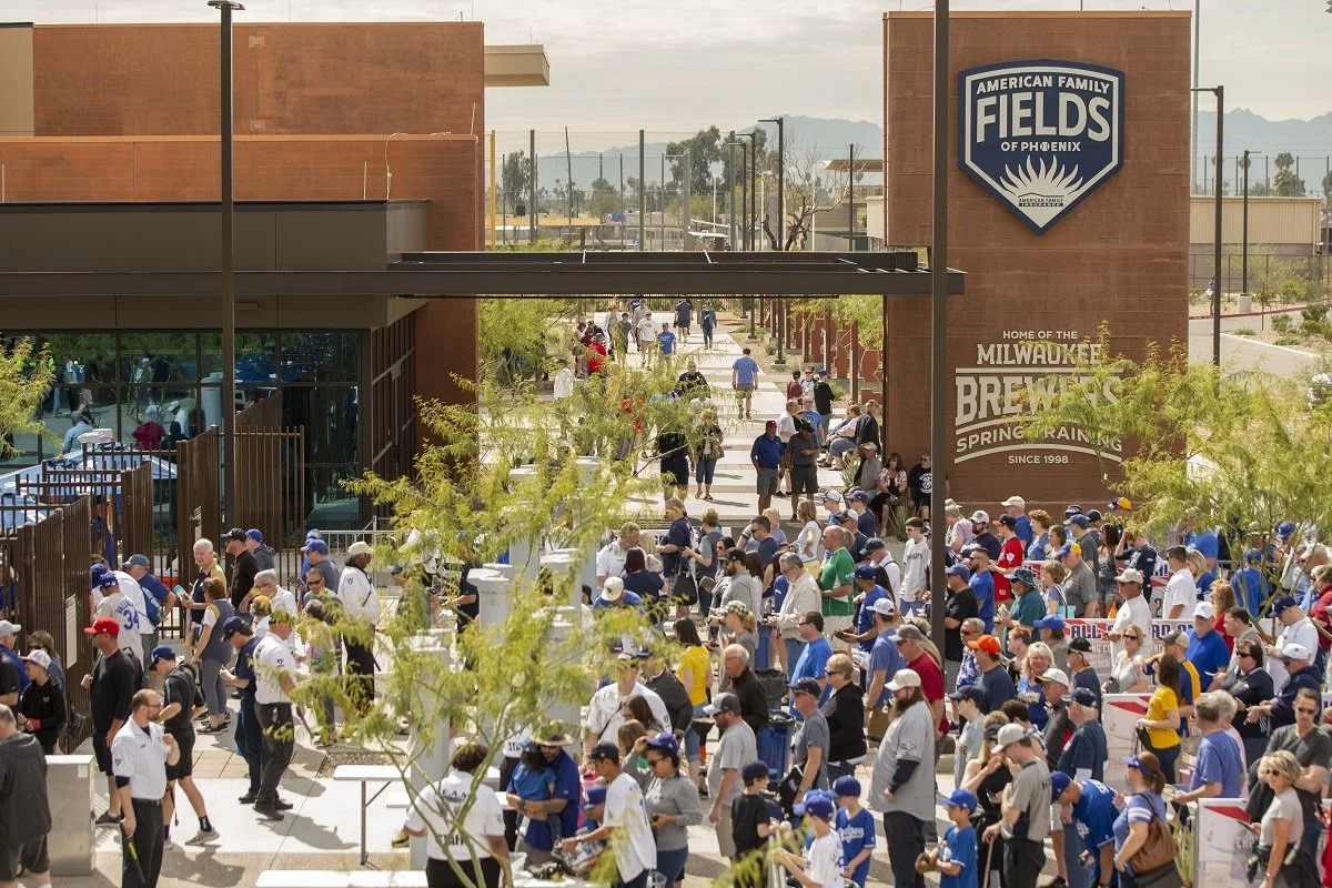 Crowds of people outside the American Family Fields of Phoenix, home of the Milwaukee Brewers spring training, on a sunny day with a large sign and buildings in the background.