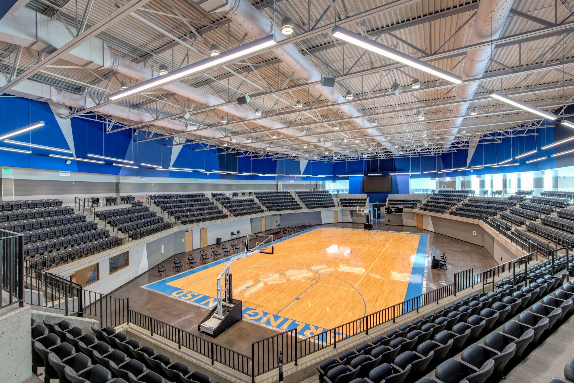 Empty indoor basketball arena with a polished court, seats on all sides, and a large electronic scoreboard at the far end.