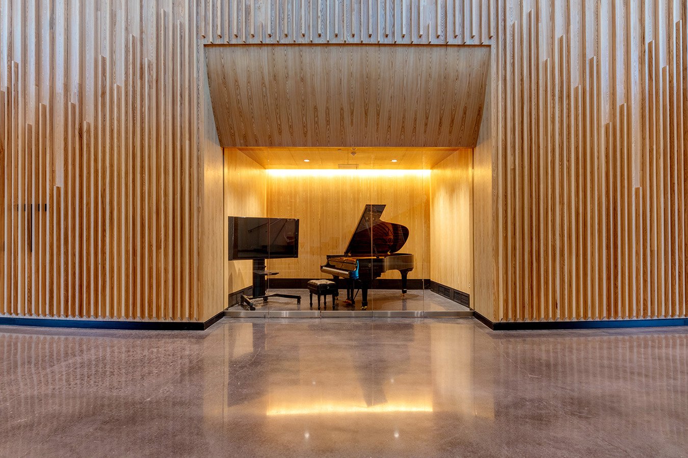 An interior view featuring a black grand piano, a flat-screen TV on a stand, and wooden panel walls with vertical slats, illuminated by warm yellow lighting.