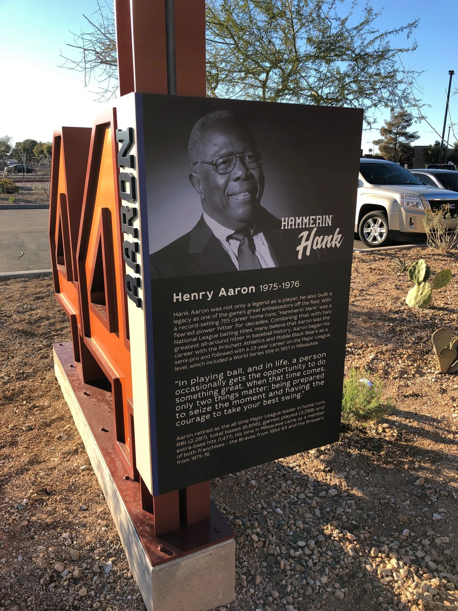 A museum EGD memorial plaque for Hank Aaron featuring a black and white photo of him, with a brief biography and quote, in a parking lot with cars and desert plants for the MLB Milwaukee Brewers. 