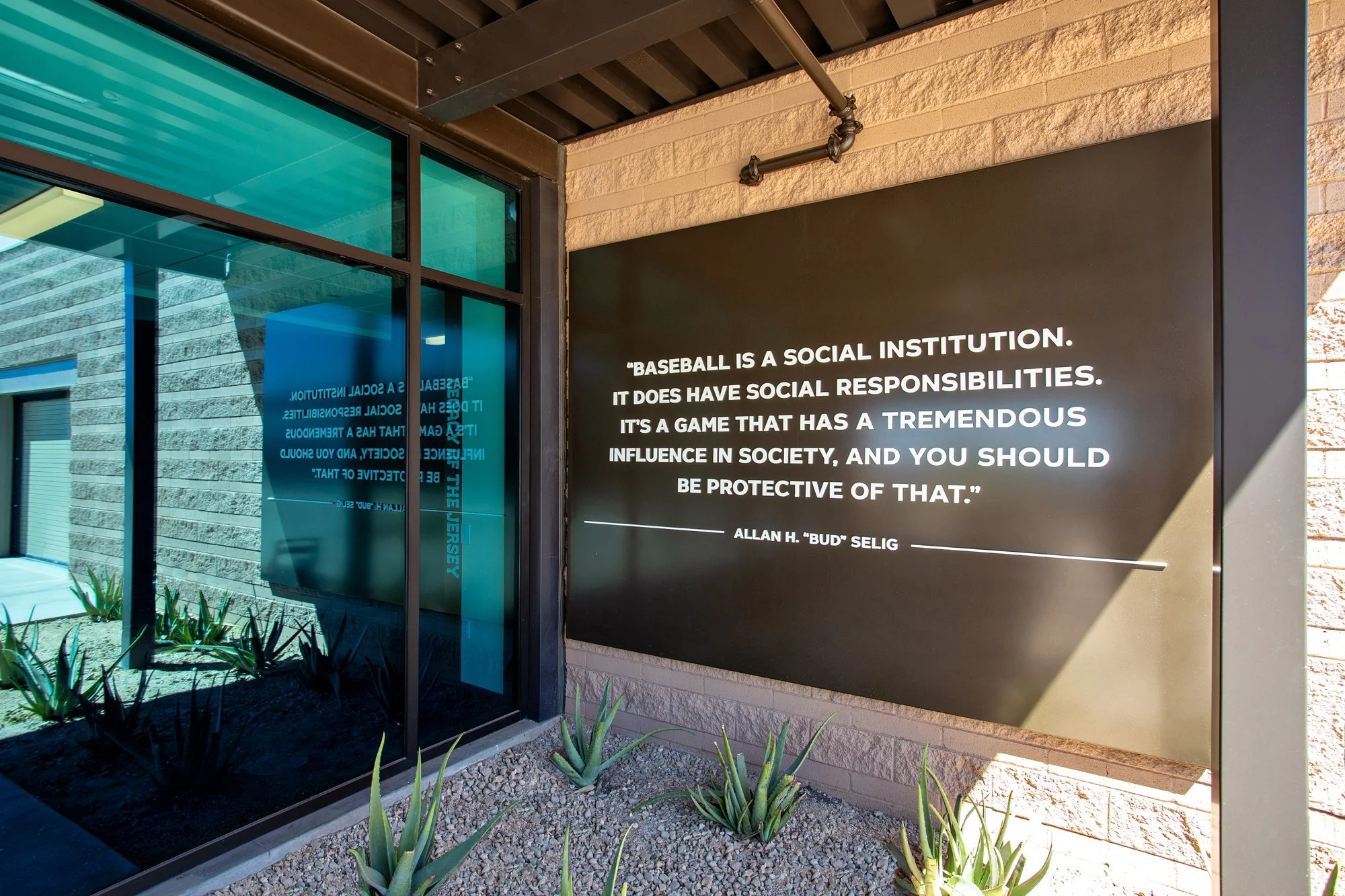 A large sign in dark brown plaque with a quote about baseball and society, attributed to Allan H. 'Bud' Selig, mounted on an exterior wall next to a glass window, with plants at the base and sunlight casting a shadow.