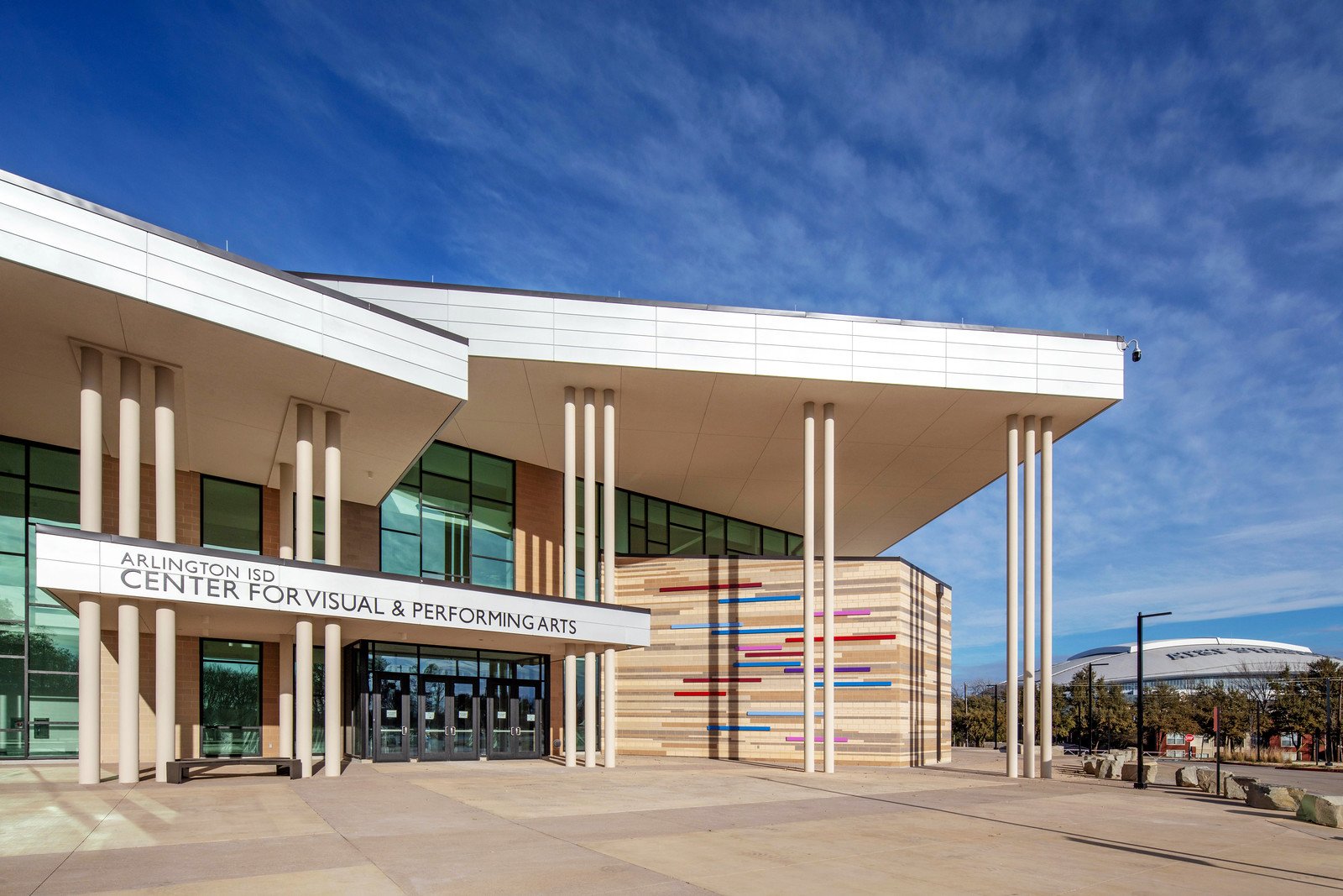 Exterior of Arlington ISD Center for Visual & Performing Arts building with modern architecture, glass windows, beige and brick walls, and a colorful wall art display under a blue sky.