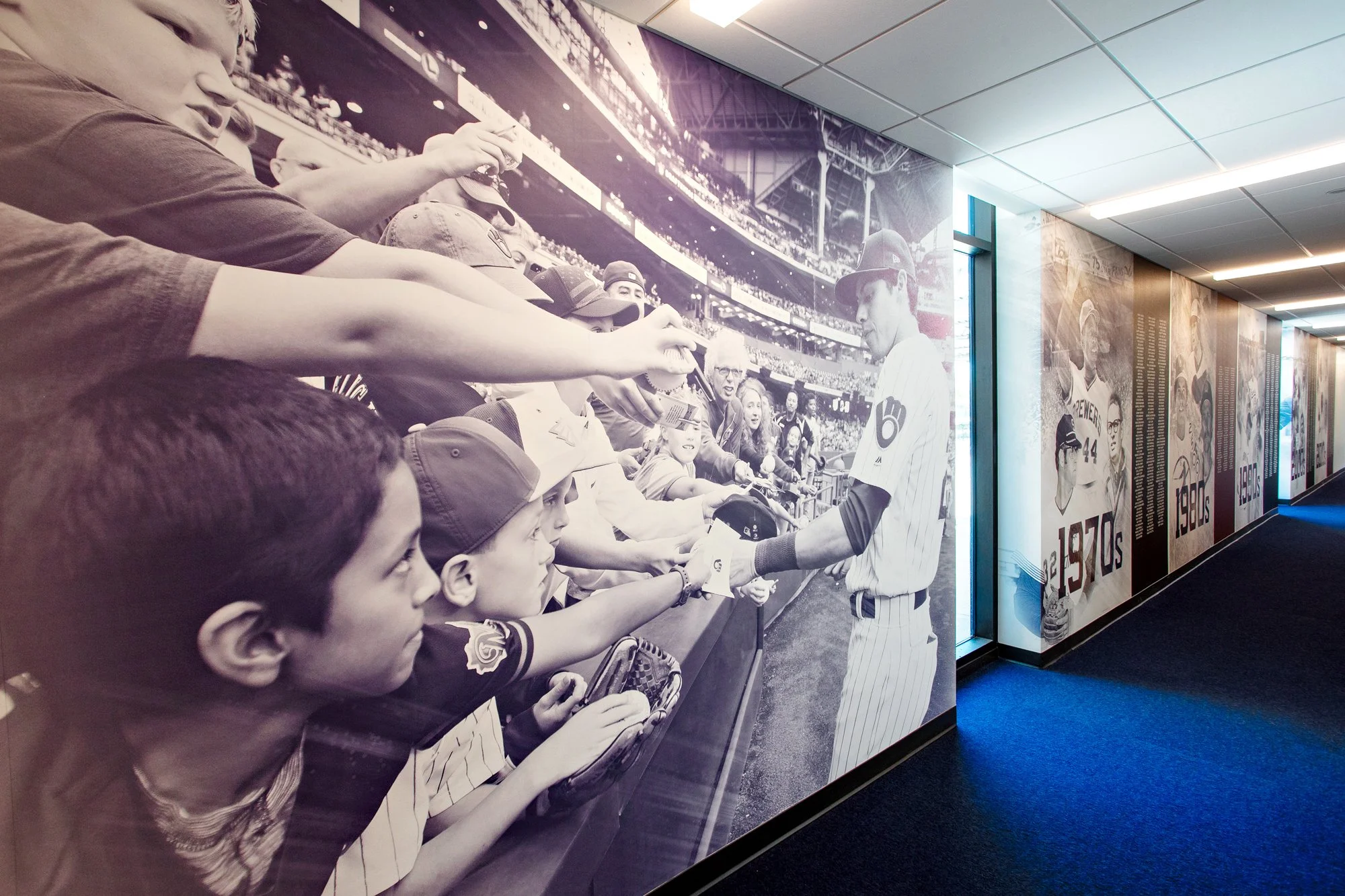 A large vinyl wall graphic of a MLB Milwaukee Brewers baseball player in uniform signing autographs for young fans through a stadium railing.