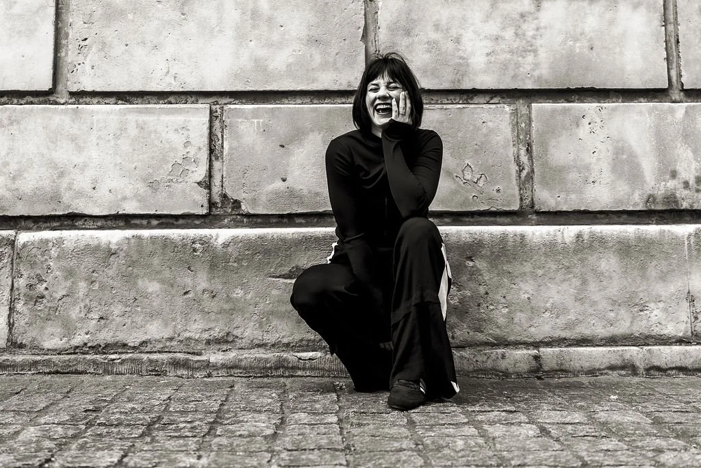 A woman with dark hair and a big smile, dressed in black, squatting and laughing against a stone brick wall, with cobblestone ground.