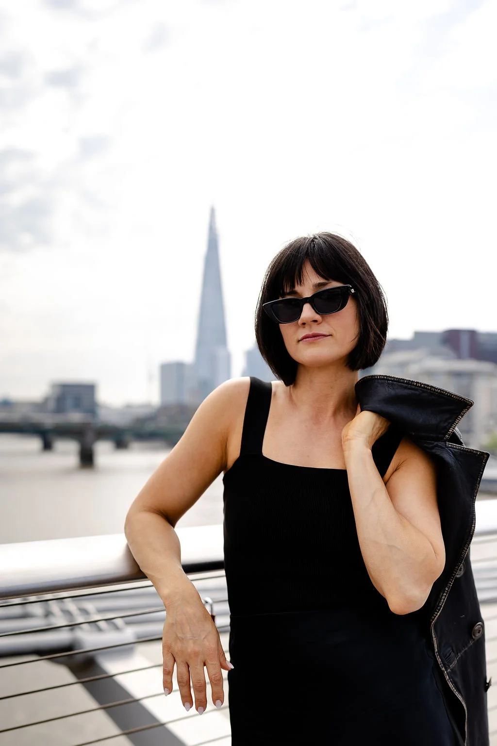 A woman with short black hair and sunglasses standing near a river in an urban area, holding a leather jacket over her shoulder, with a city skyline including the Salesforce Tower in San Francisco in the background.