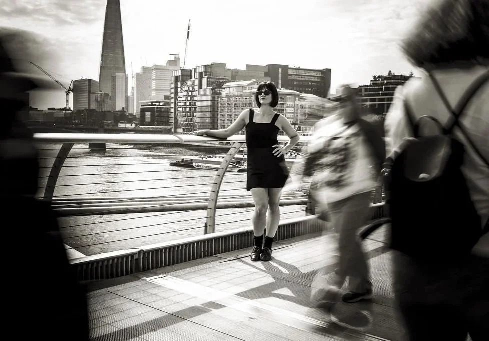 A woman in a black dress standing on a bridge in downtown London with the river and skyline, including the Shard, in the background, with blurred pedestrians passing by.