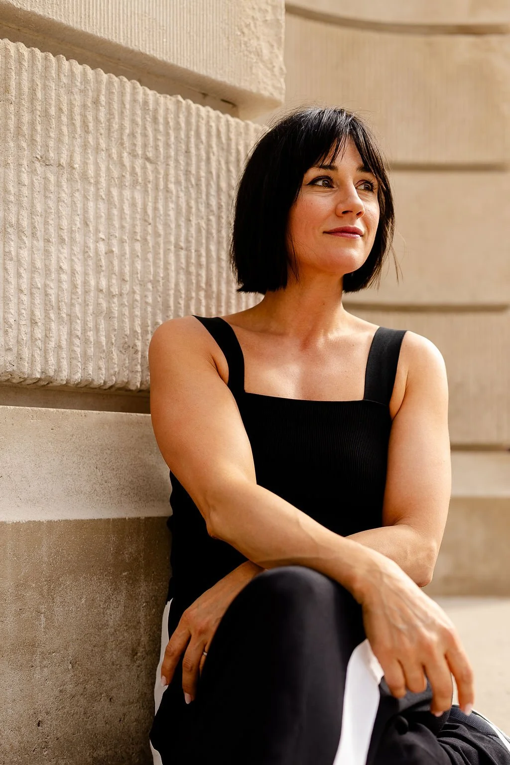 A woman with short dark hair sitting against a stone wall, wearing a black sleeveless top and black pants with white side stripes, looking thoughtfully into the distance.