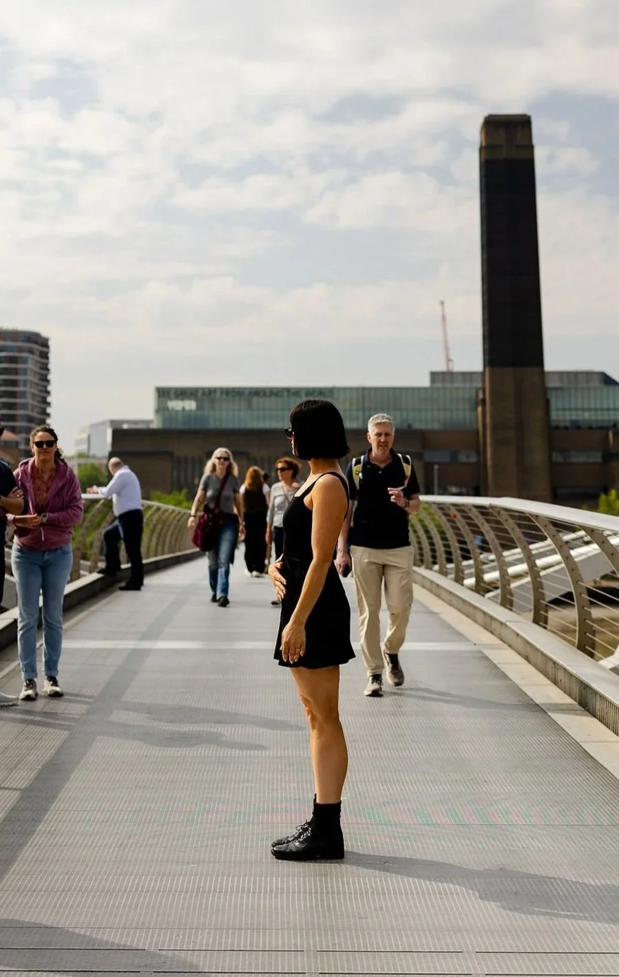 A woman with short black hair in a black dress and boots stands on a pedestrian bridge, with other people walking and talking around her, in an urban setting with modern buildings and a tall chimney in the background.