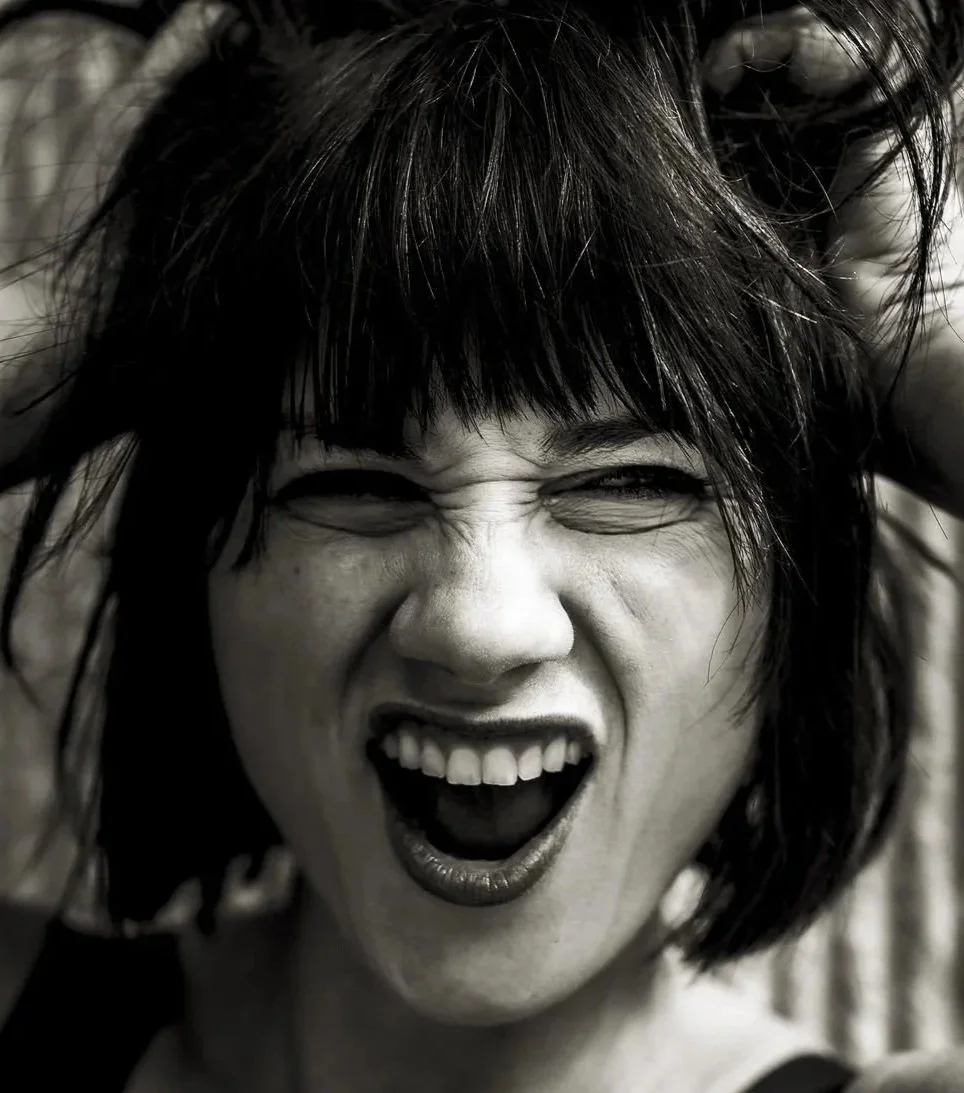 A woman with dark, messy hair is making a fierce, angry facial expression, squinting her eyes and baring her teeth.
