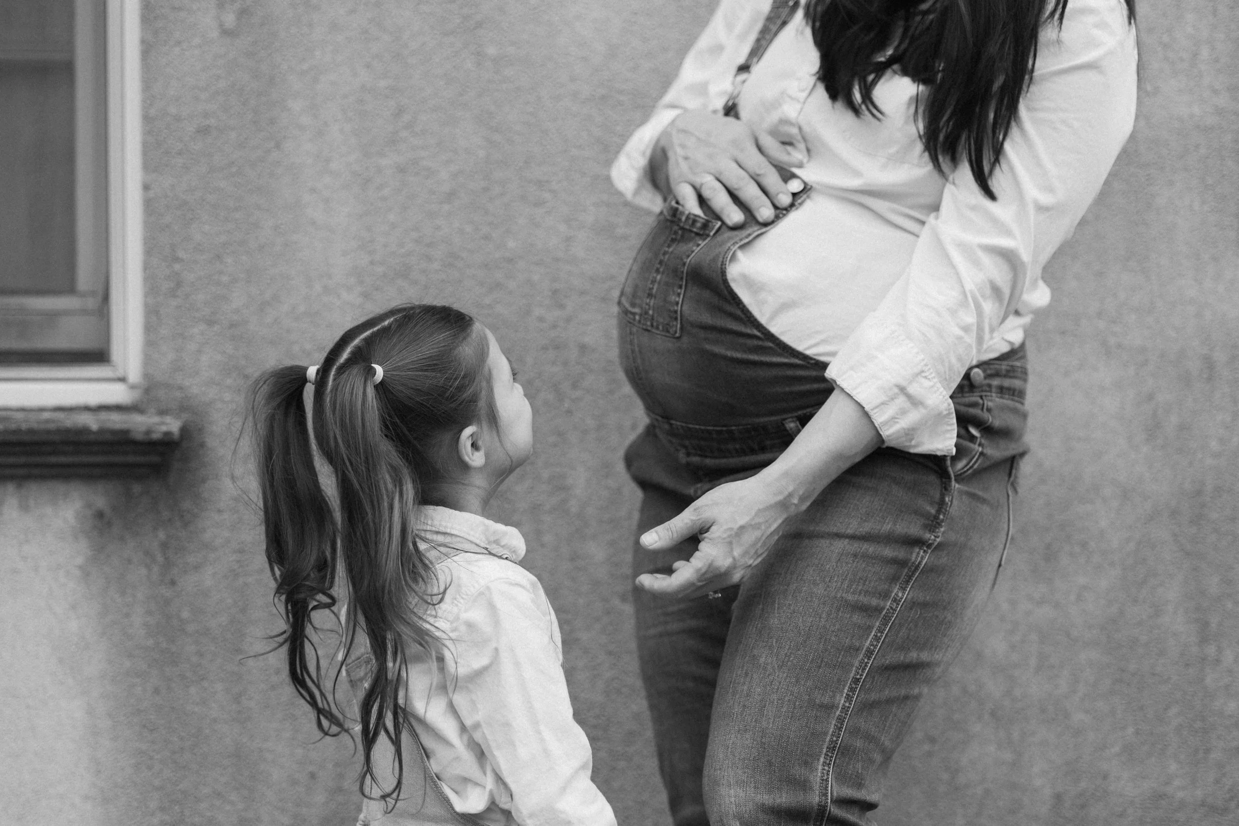 A woman bending down towards a young girl with long hair tied in pigtails, inside a room with a textured wall and a window.