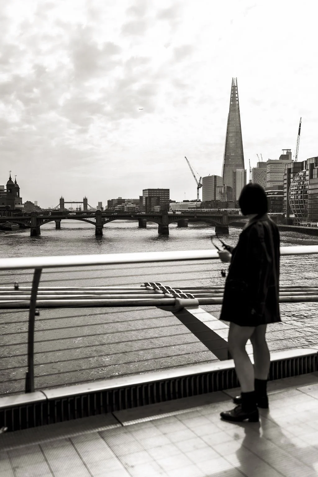 A black-and-white photo of a city skyline with the Shard building, Thames River, and Tower Bridge visible in London. A person is standing by the railing, looking at a phone.