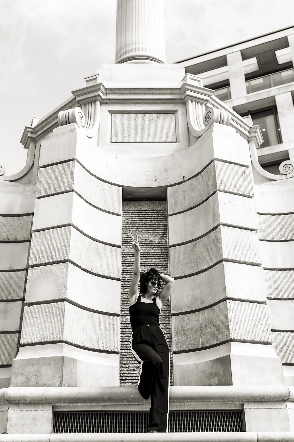 Woman in black outfit and sunglasses posing against a historic stone building with classical architectural details.