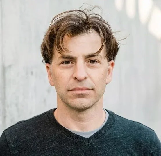 Headshot of founder of Group Design Build, M. Tagore Hernandez a man with medium-length brown hair, wearing a dark sweater, standing outdoors near a white wooden wall.