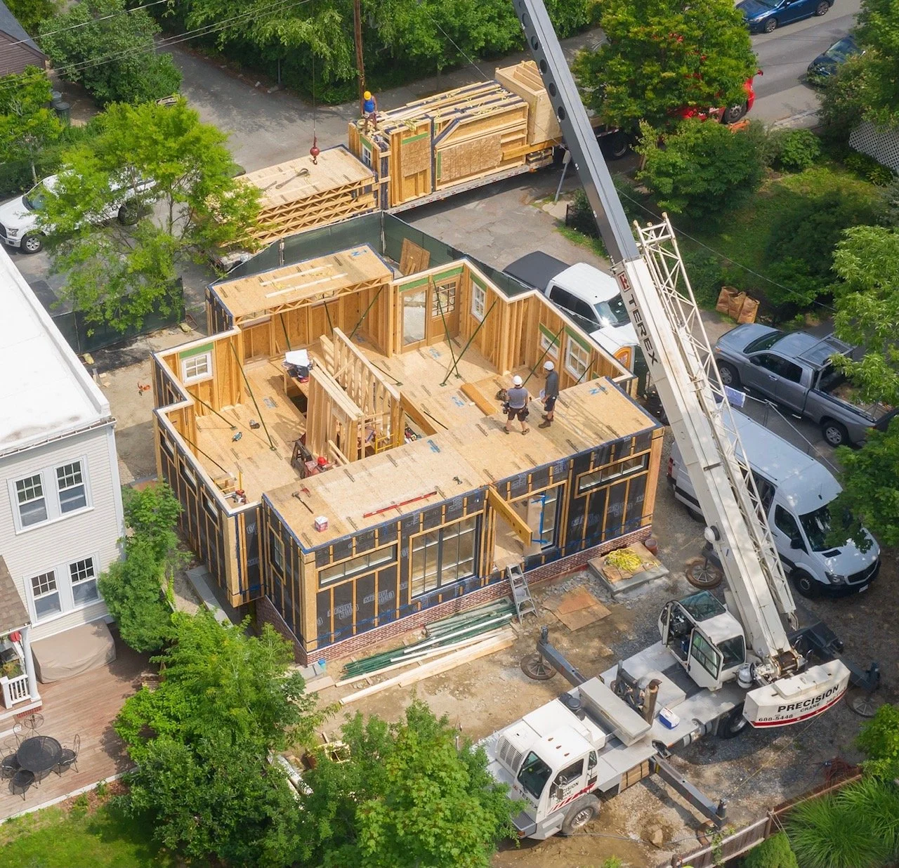 A house under construction with wooden framing, construction workers, and a crane lifting materials. Nearby are parked cars and a neighboring house with a small patio.