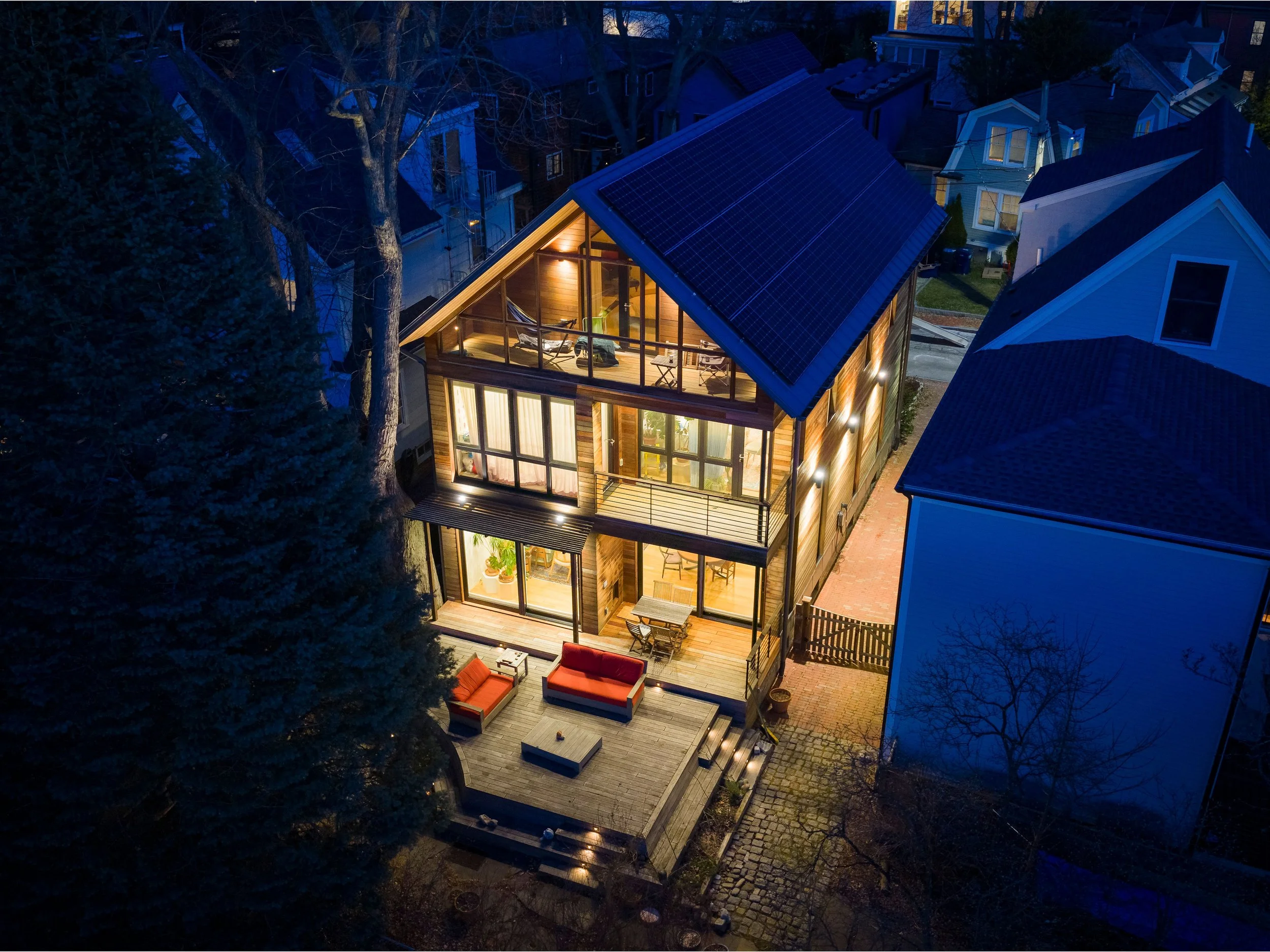 A nightly view of a modern house with large glass windows and a balcony, illuminated from inside, featuring solar panels on the roof. The house is in a neighborhood with other houses visible nearby, surrounded by leafless trees.