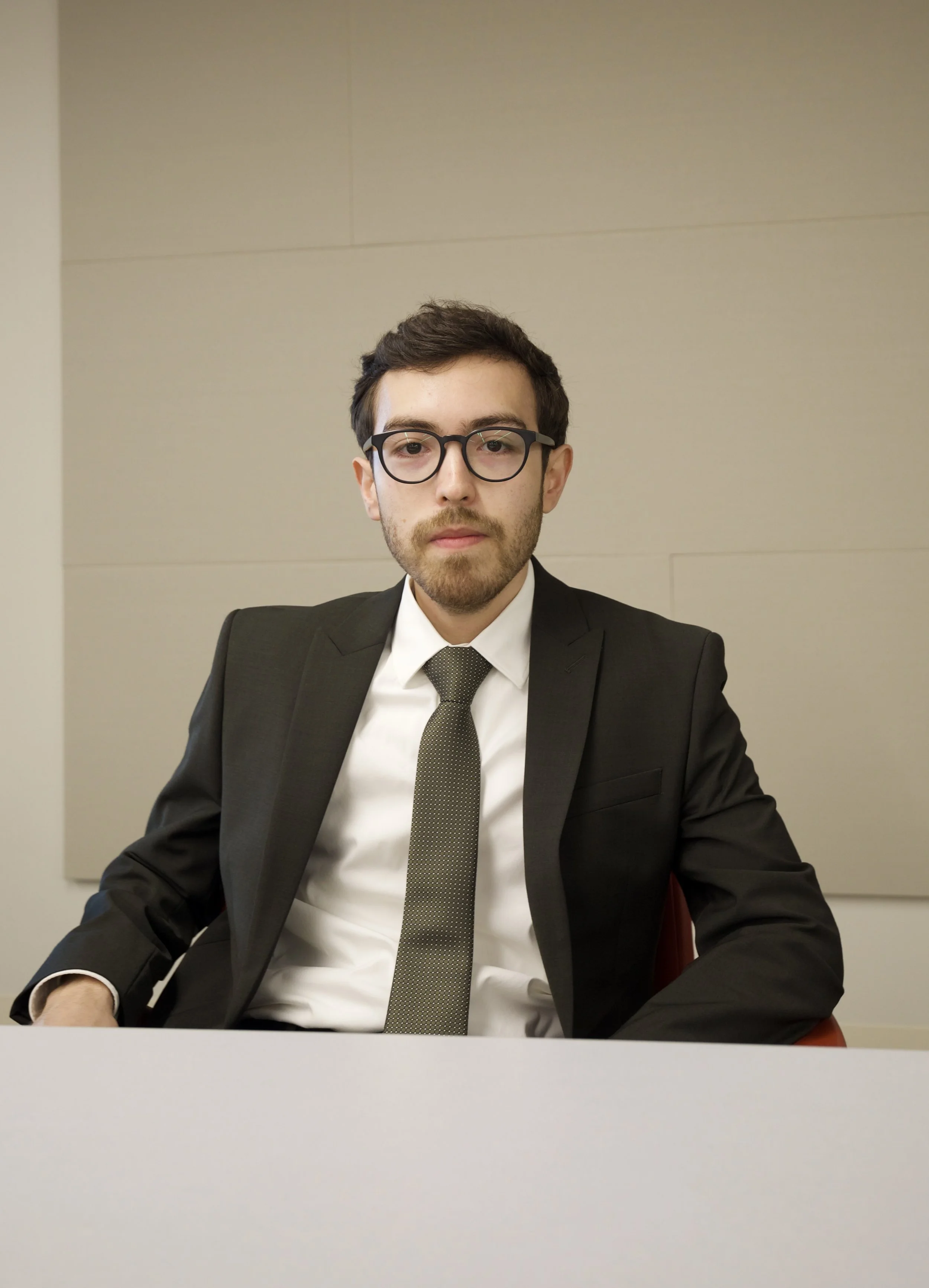 A young man in a business suit and glasses sitting at a table in an office with a plain wall.