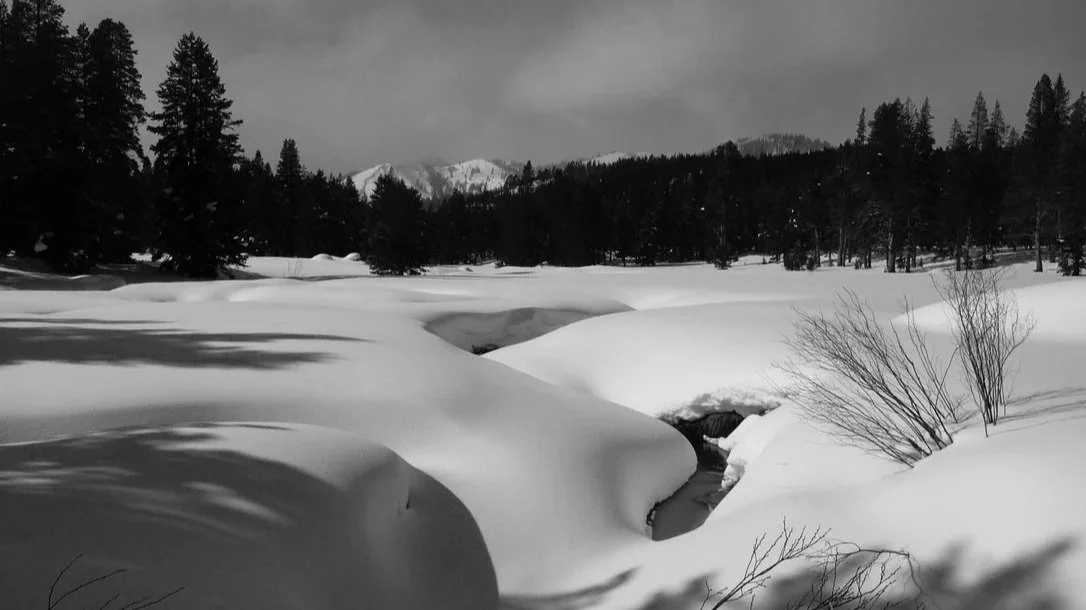 Snow-covered landscape with a small partially frozen stream, trees, and mountains in the background under cloudy sky.