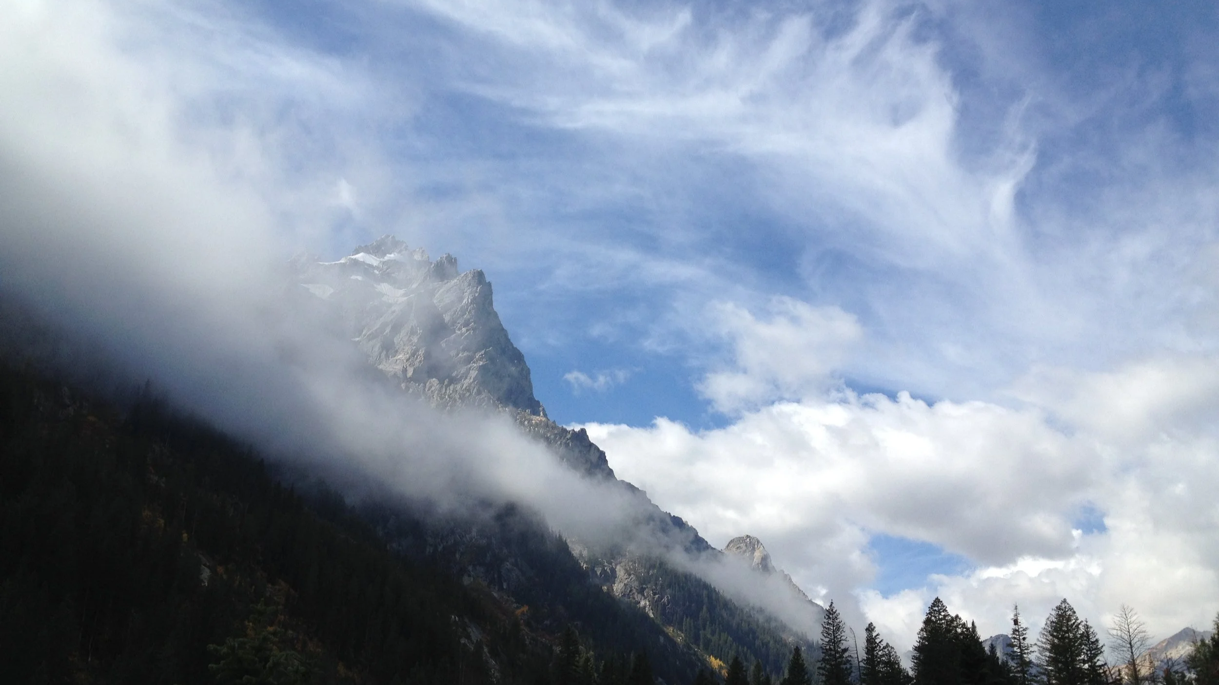 A mountain landscape with a snow-capped peak, surrounded by clouds and a blue sky with wispy clouds. Forested slopes are in the foreground.