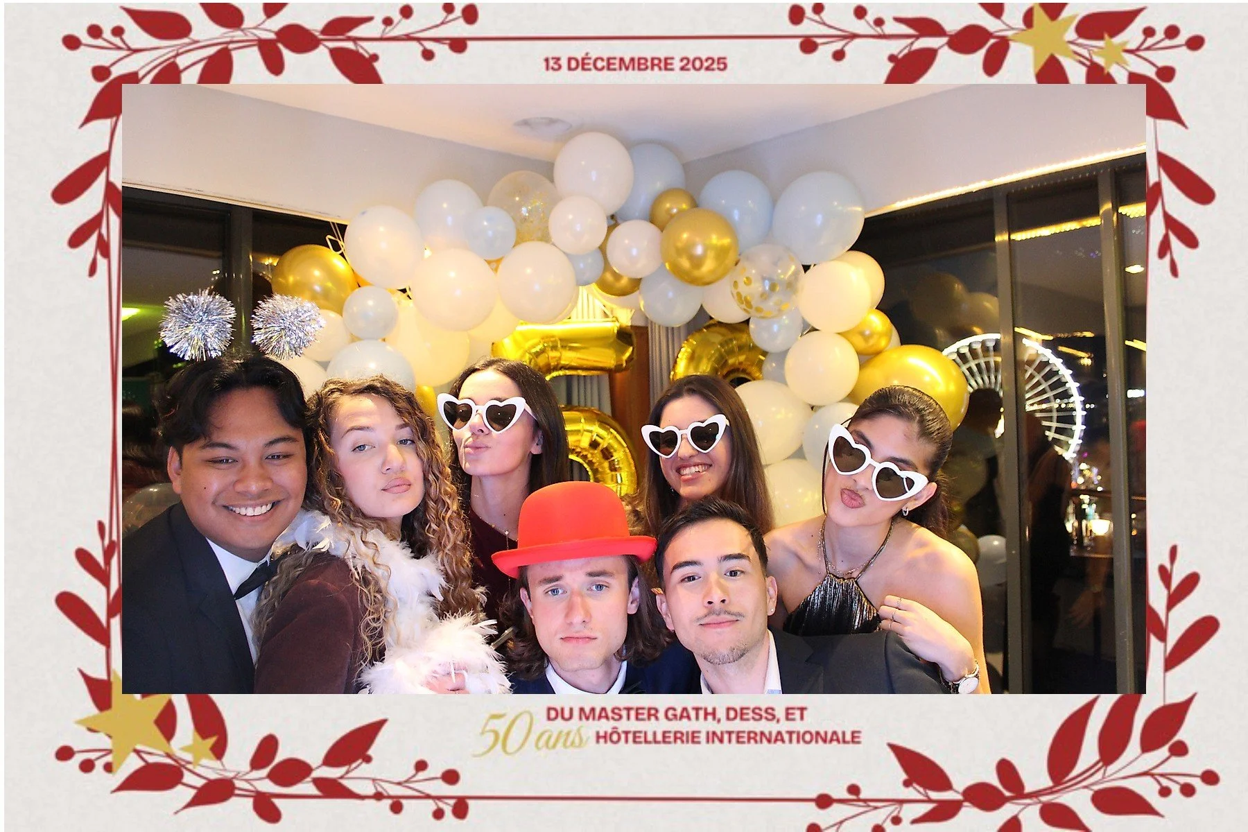 Groupe de personnes célébrant un événement avec des décorations de ballons blancs et or, portant des accessoires amusants comme des lunettes en forme de cœur et un chapeau rouge, dans une salle décorée pour fêter les 50 ans de l'Hôtellerie Internatio