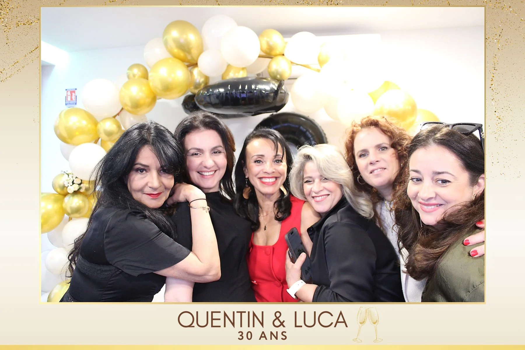 Un groupe de six femmes souriantes posant lors d'une célébration de 30 ans pour Quentin et Luca, avec un décor de ballons dorés, blancs et noirs en arrière-plan.