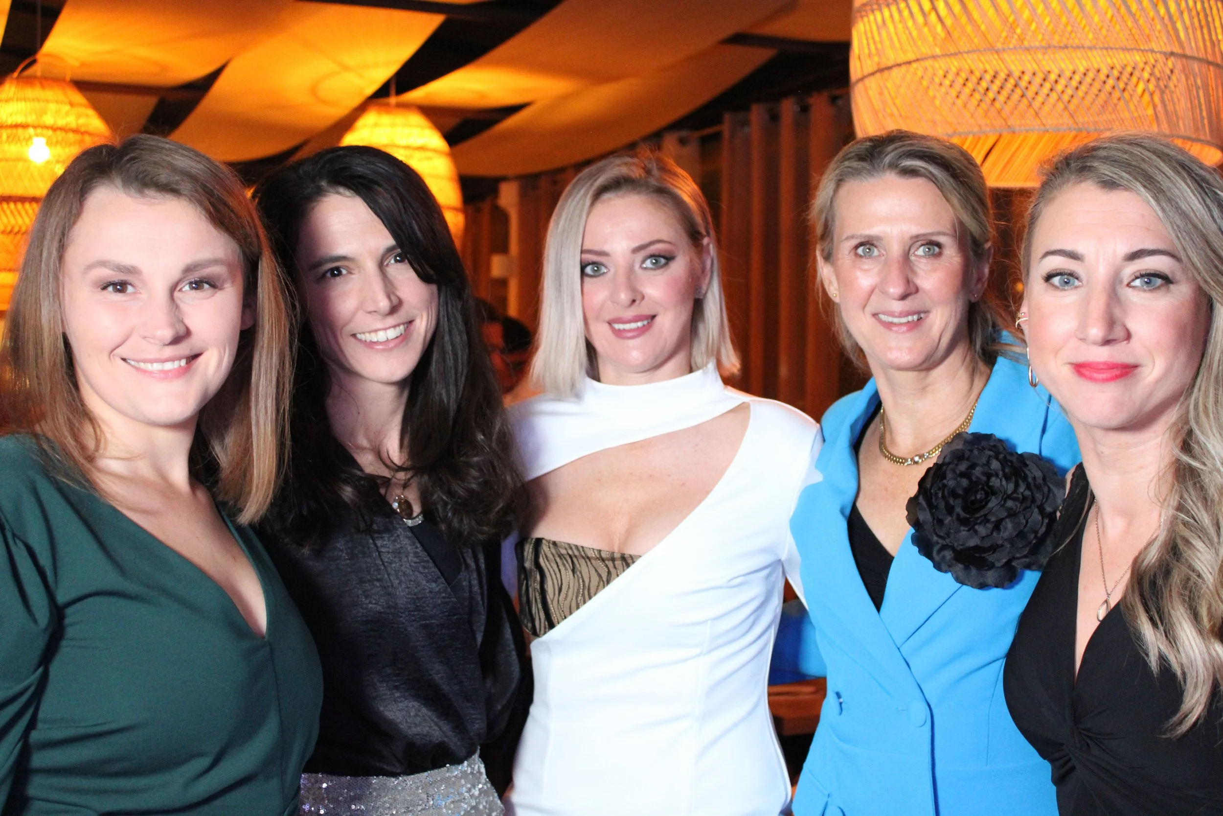 Cinq femmes souriantes posant pour une photo lors d'un événement social dans un lieu avec décoration chaleureuse et éclairage tamisé.