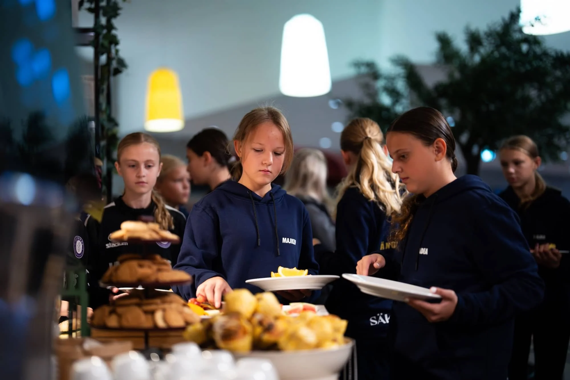 Ungdomar i kö vid ett buffébord med bakverk och frukt i en inomhusmiljö.