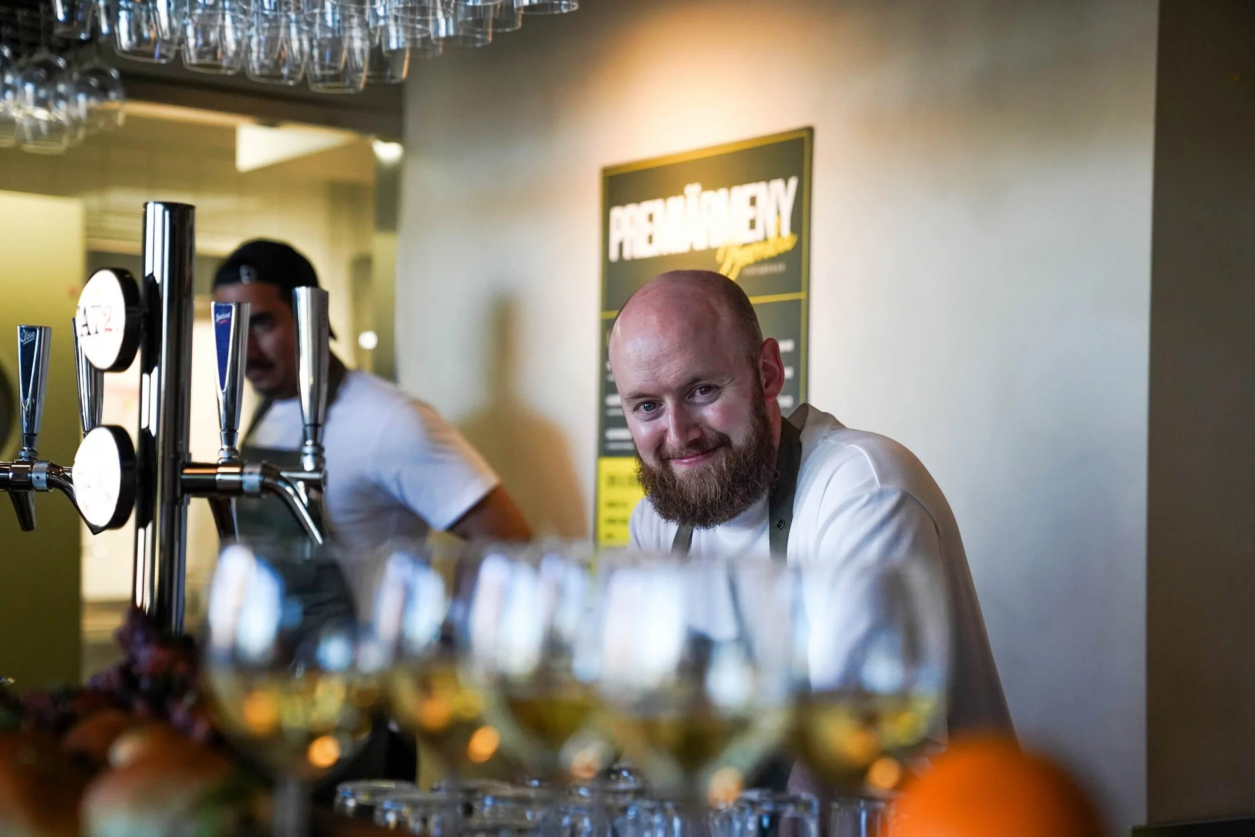 Två bartenderar serverar drinkar på en bar, en av dem tittar mot kameran.