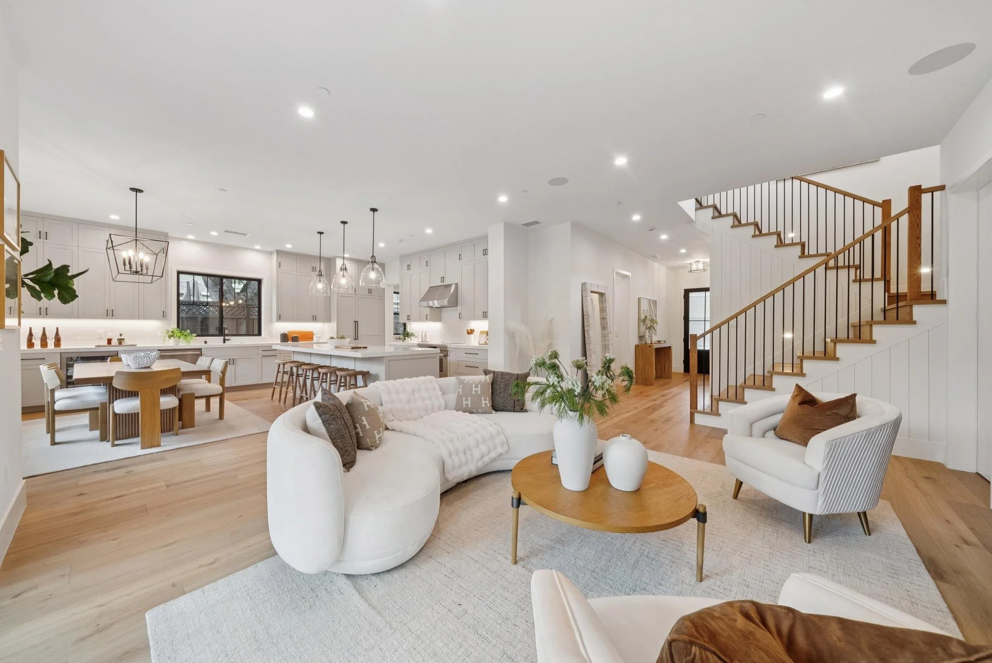 Open-concept living room and kitchen with white cabinets, wooden floors, and modern furniture including a white sectional sofa, armchair, and wooden dining table, with staircase in the background.