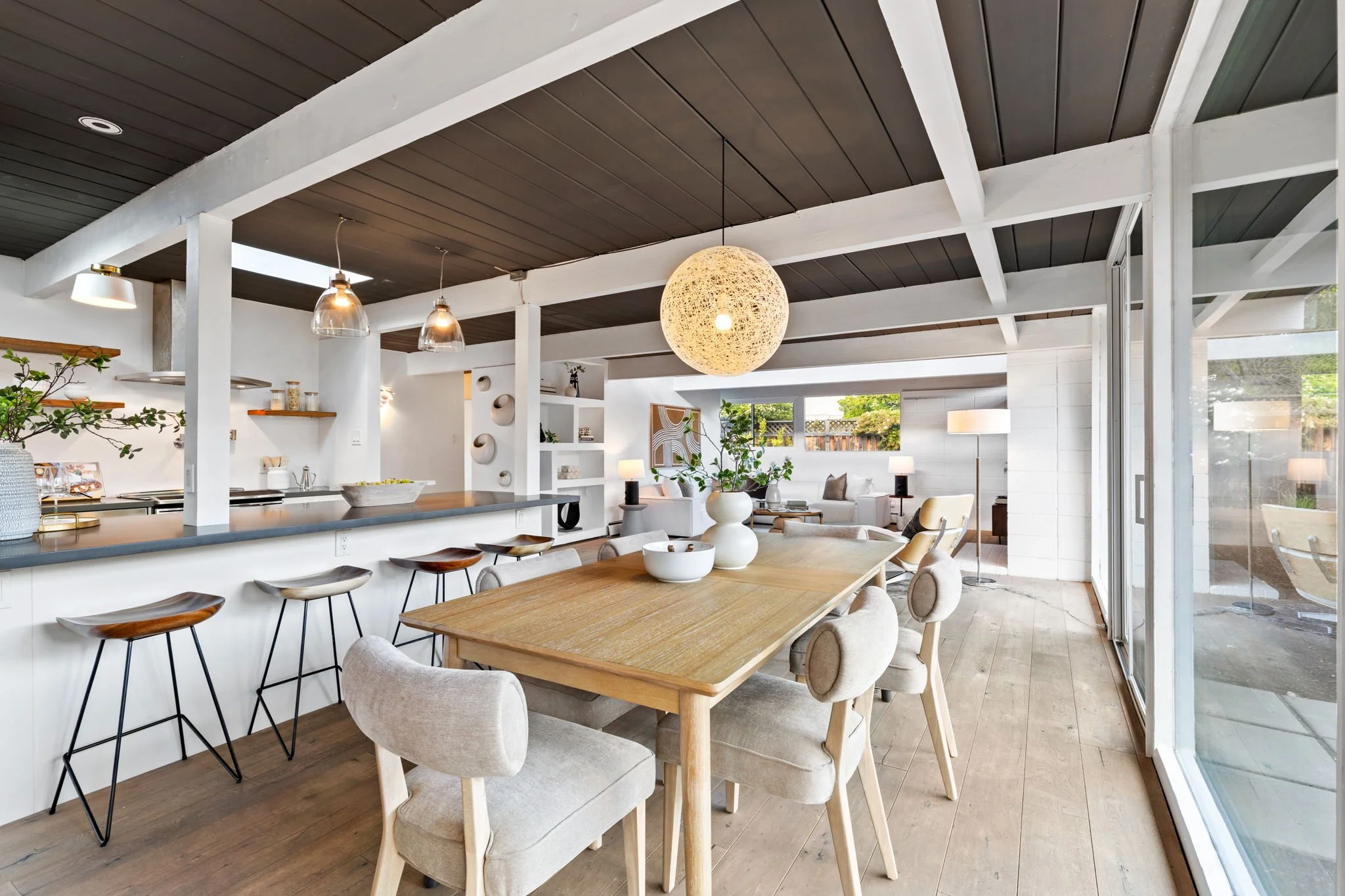 Open-concept living and dining area with a wooden dining table, beige upholstered chairs, a white hanging pendant light, a living room with white sofas, armchairs, and side tables, large windows with a view outside, and a modern, minimalist decor style.