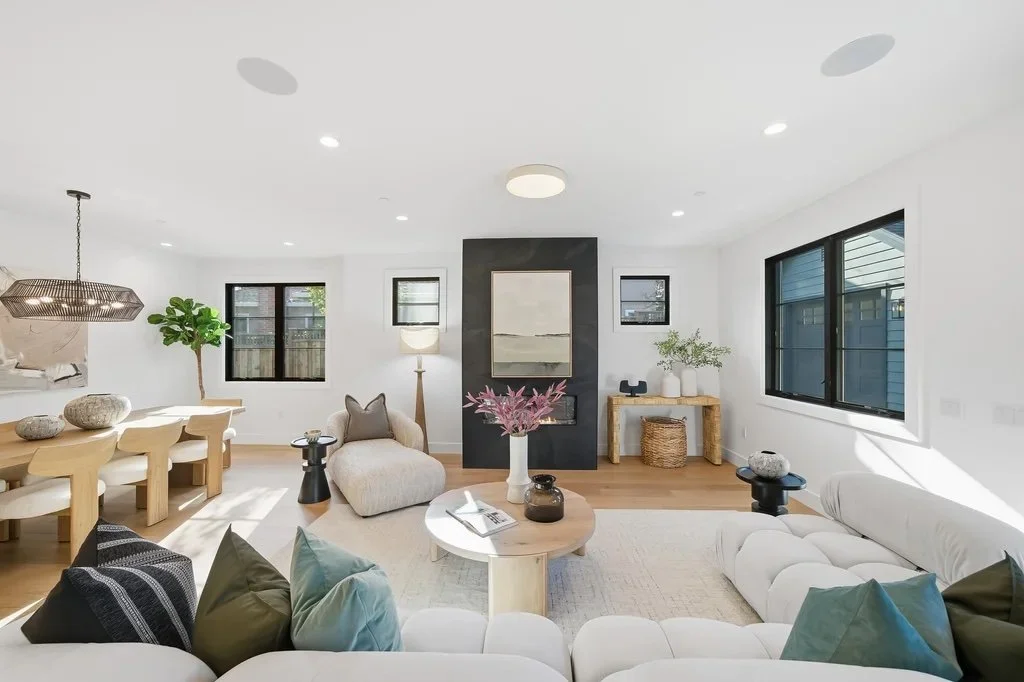 Modern living room with white walls, large windows, and minimalist decor including a white couch with throw pillows, a wooden coffee table with vases, a cozy armchair, and a fireplace with a black accent wall.