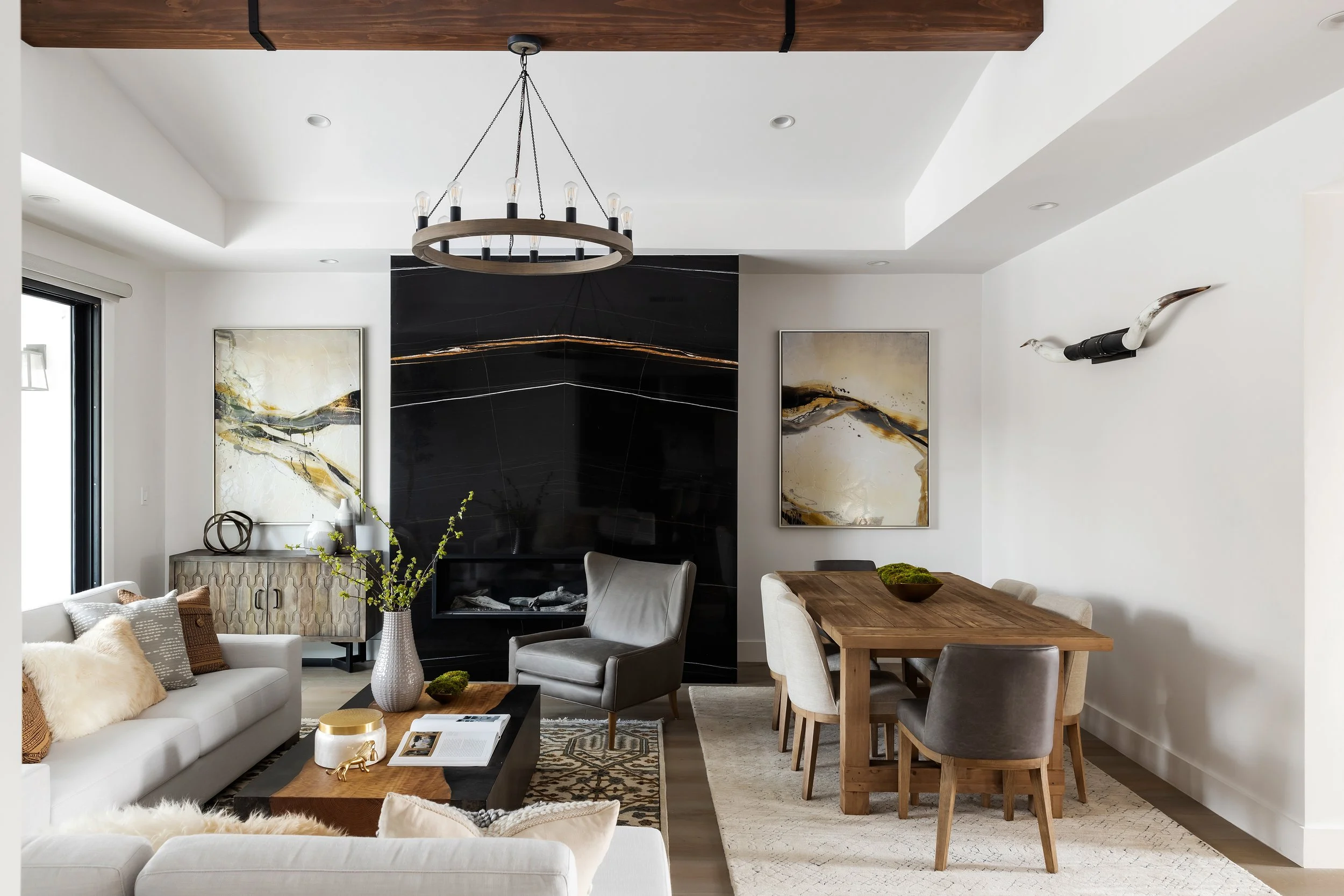 Modern living room with white and neutral tones, featuring a white sofa with textured and furry pillows, a black coffee table with decor, artworks on the walls, a large vase with branches, a gray armchair, a wooden dining table with upholstered chairs, and a black fireplace with a black marble surround. Elegant lighting fixtures and decorative wall art complete the space.