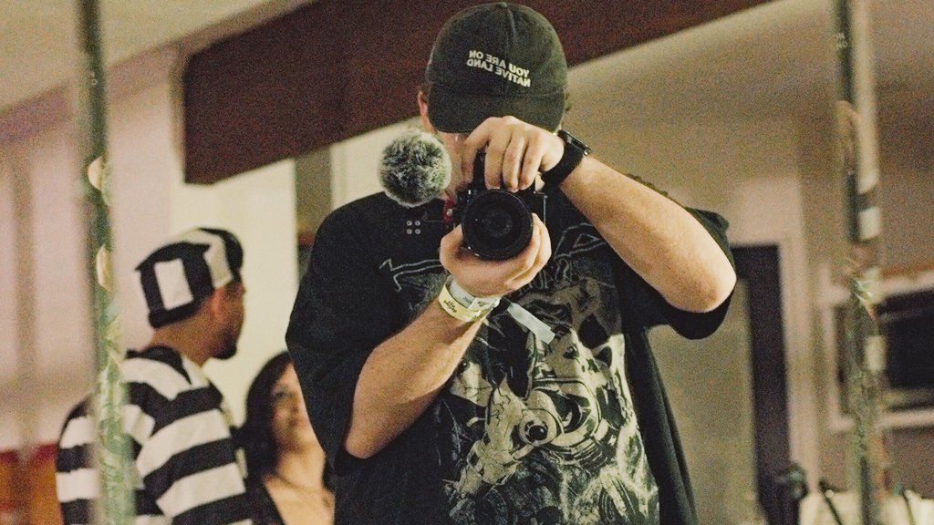 Person taking a photo in a mirror, wearing a dark cap and a black T-shirt with a graphic print, holding a camera with a microphone attached, in an indoor setting with people in the background.
