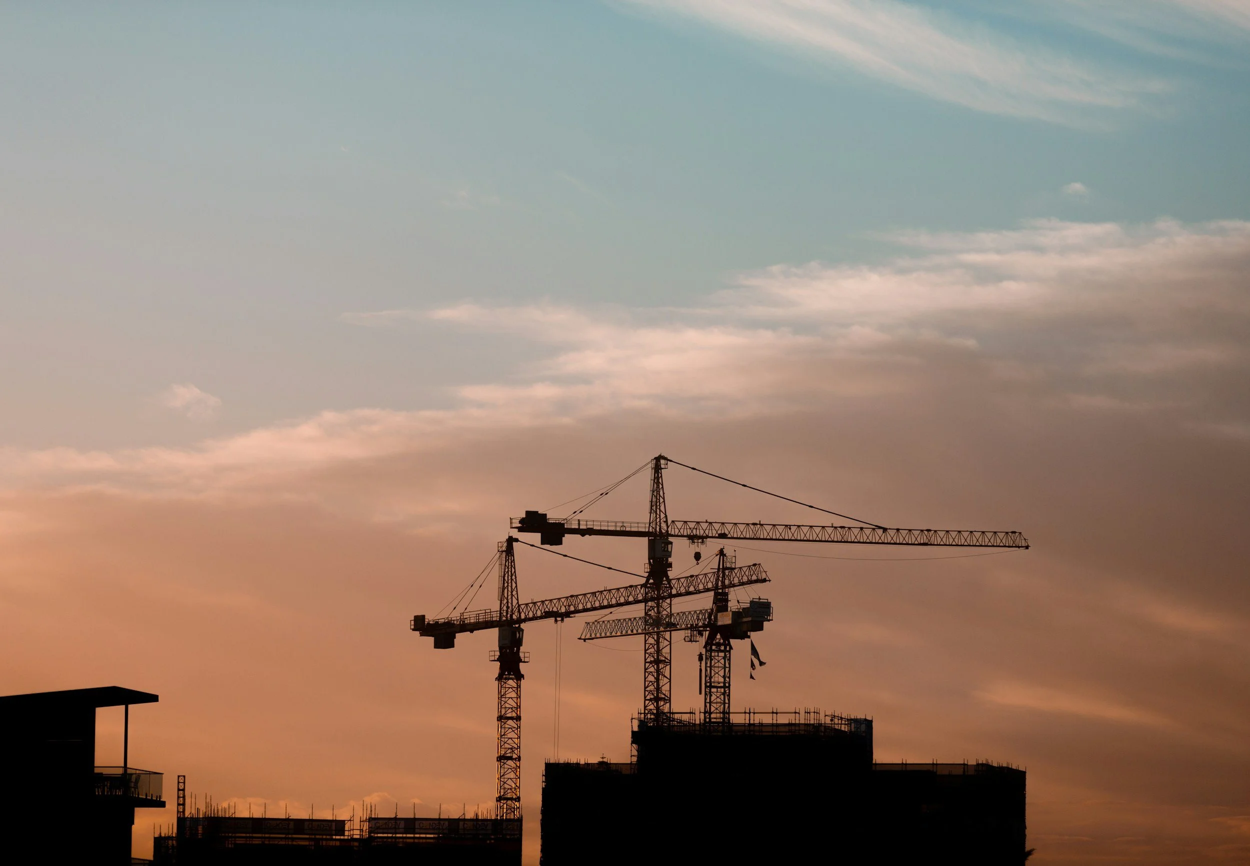 Construction cranes on a building site during sunset or sunrise with a partly cloudy sky.