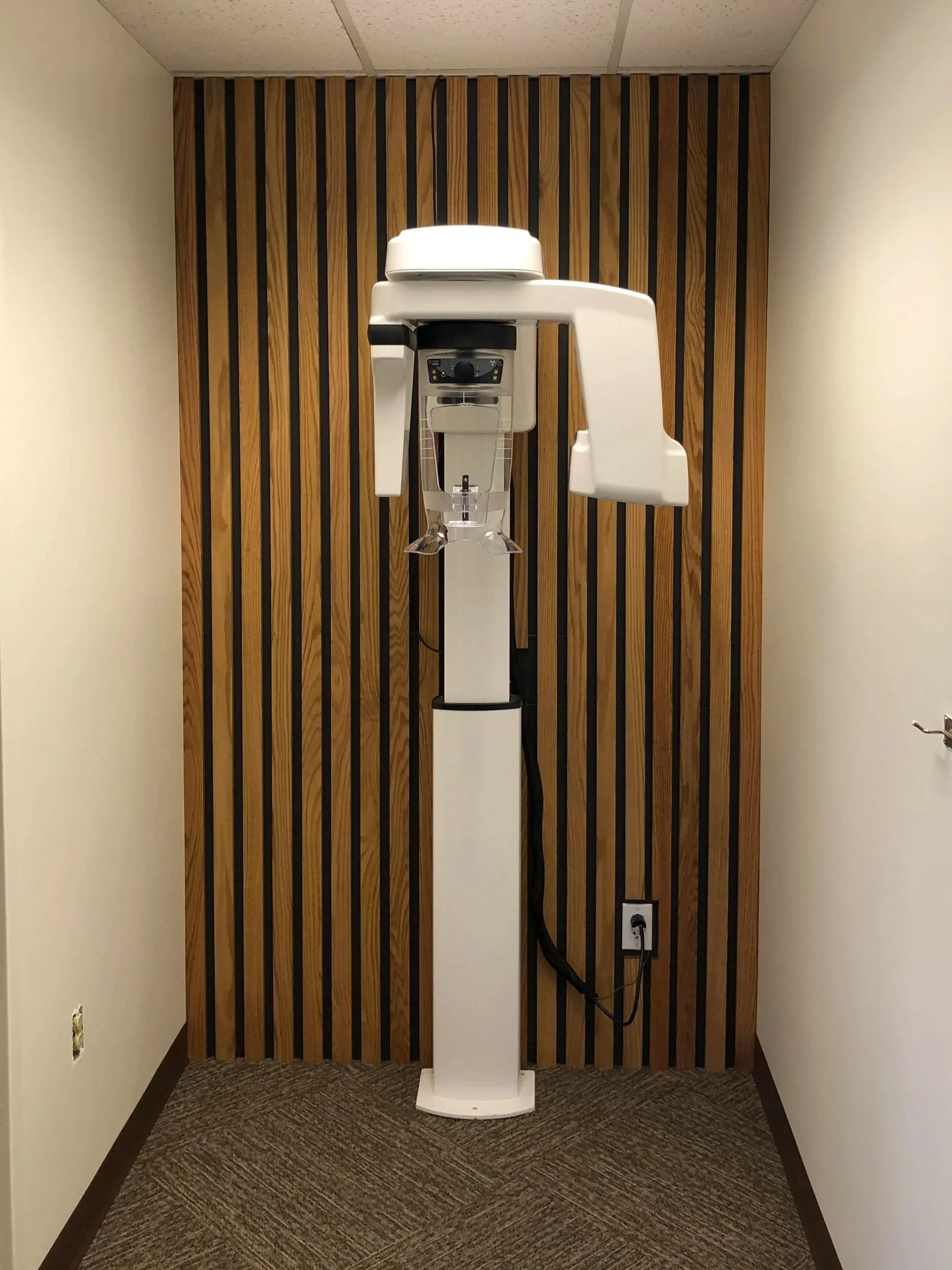 A standing X-ray machine in a room with wooden paneling on the back wall and beige walls on the sides, plugged into an electrical outlet on the right wall.